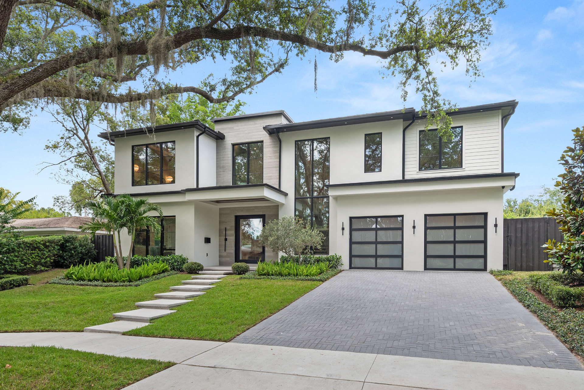 Front elevation of Munster: a two-storey white stucco and stone residence with black-framed floor-to-ceiling windows, glass garage doors, floating concrete steps and layered planting beneath a live oak.