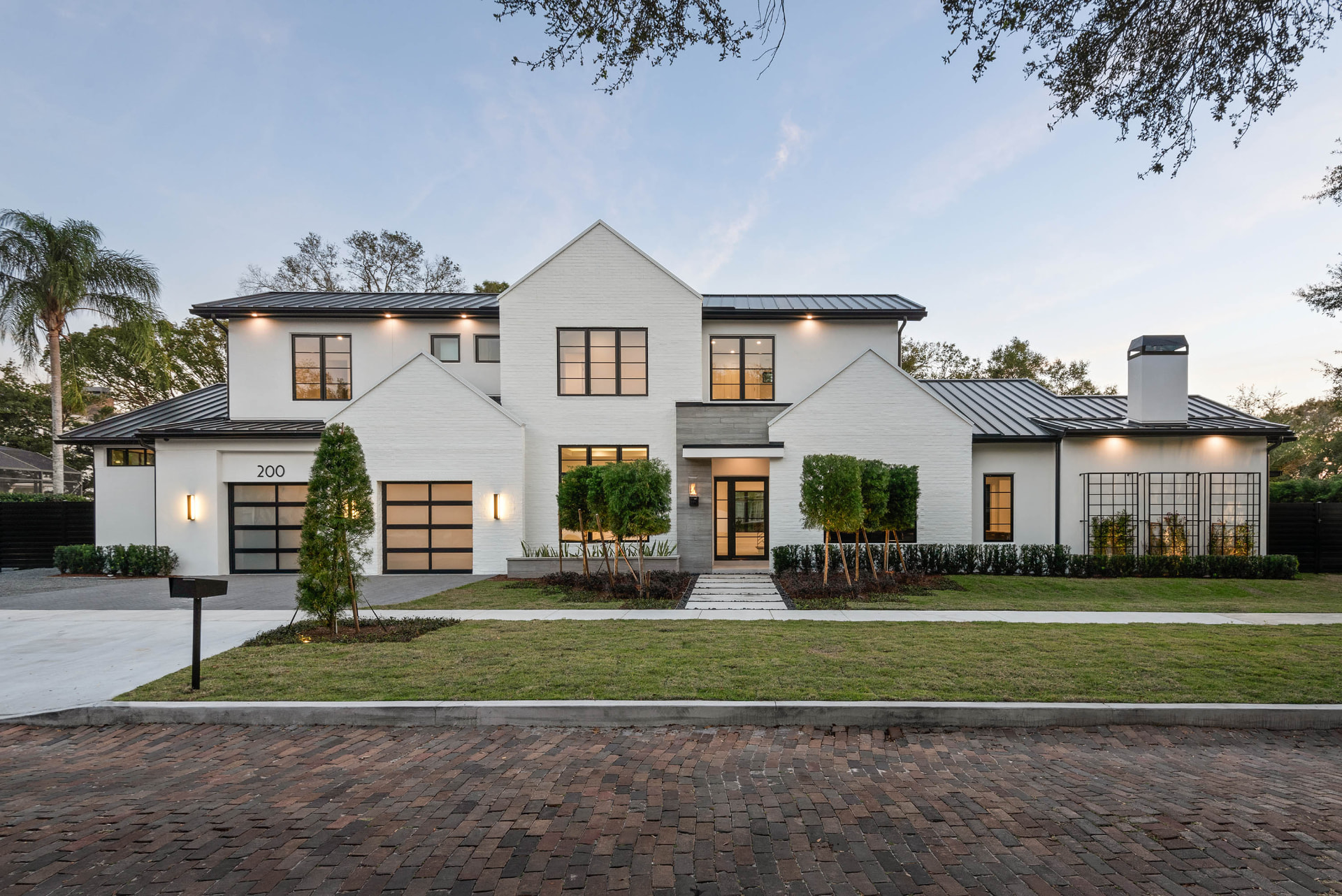 Front elevation of Oakwood: a white painted brick residence with steep gables, a black standing-seam metal roof, black-framed windows and glass garage doors, a recessed stone entry, clipped hedges and slender topiary above a brick paver street.