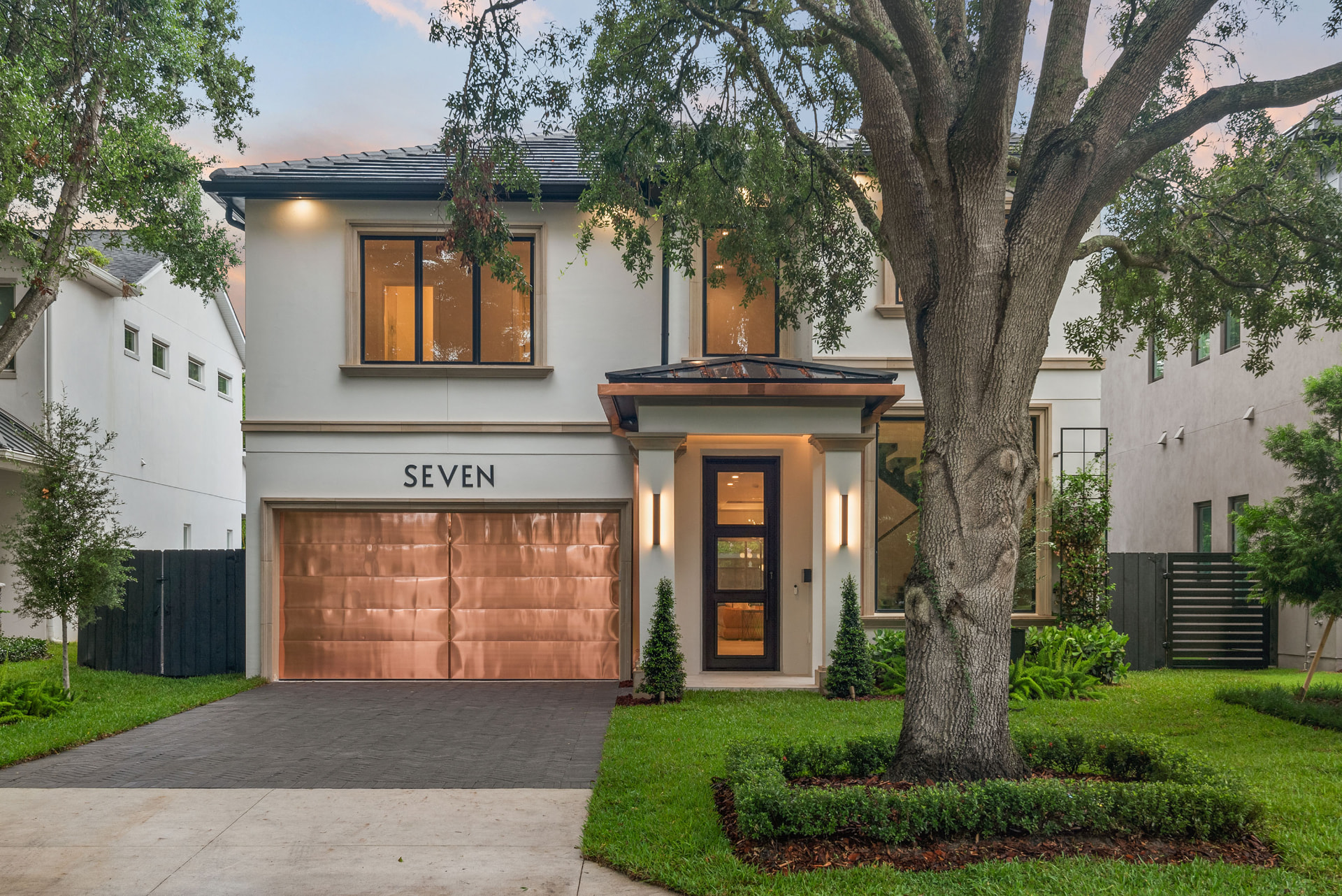 Front elevation of New Hampshire at dusk: a two-storey stucco residence with a copper garage door and copper entry canopy, warm lit interiors and a mature oak in the front lawn.