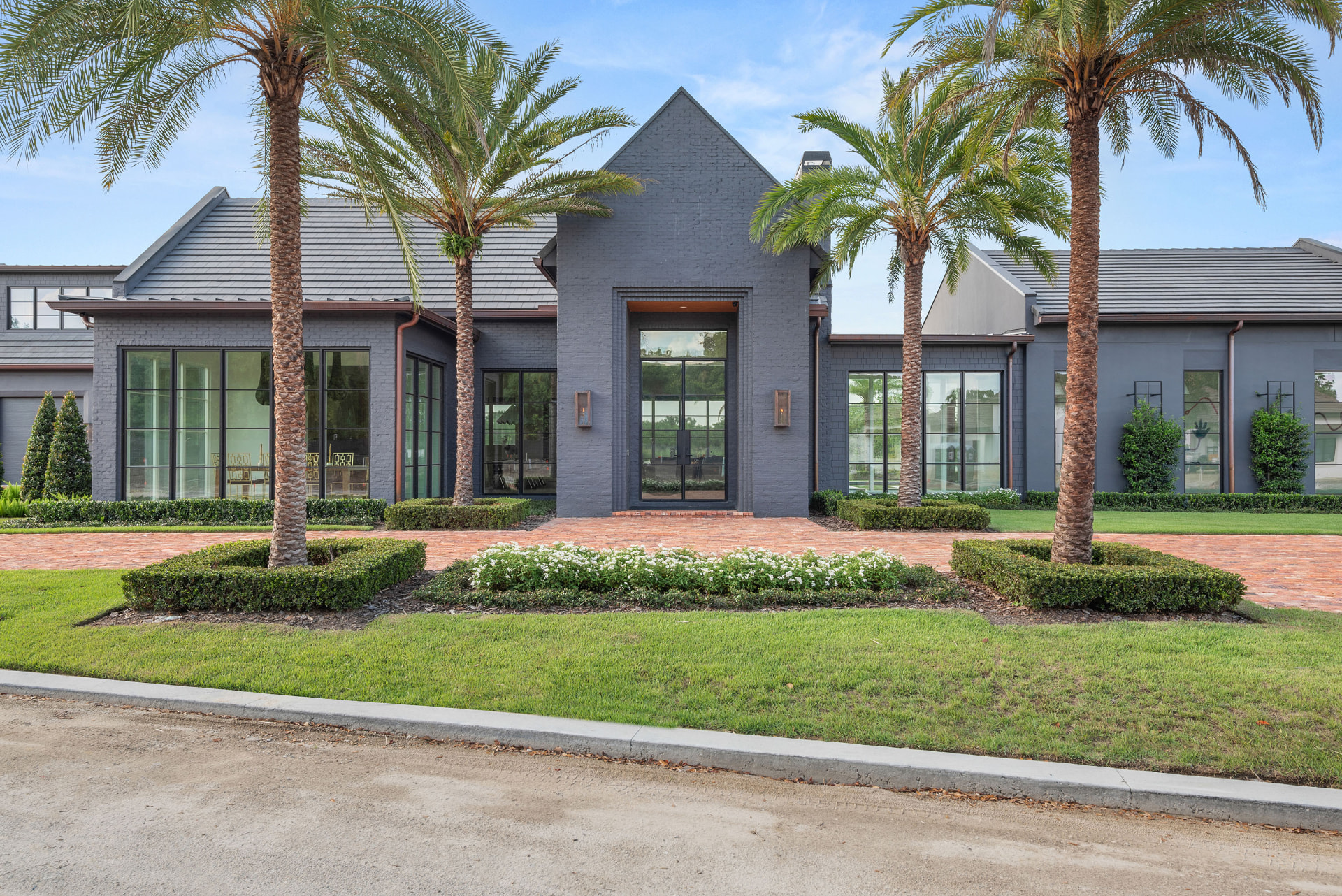 Front elevation of Manor: a dark grey brick residence with a steep gabled entry, tall black-framed windows, copper downspouts, mature palms and clipped hedges along a brick paver drive.