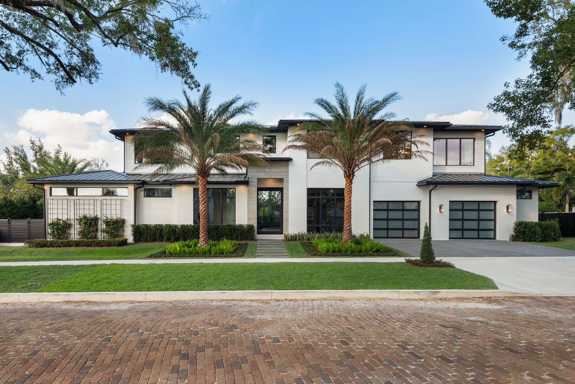 Front elevation of Rockwood: a two-storey white painted brick residence with black standing-seam roofs, black-framed glass garage doors, two tall date palms and clipped hedges above a brick paver street.