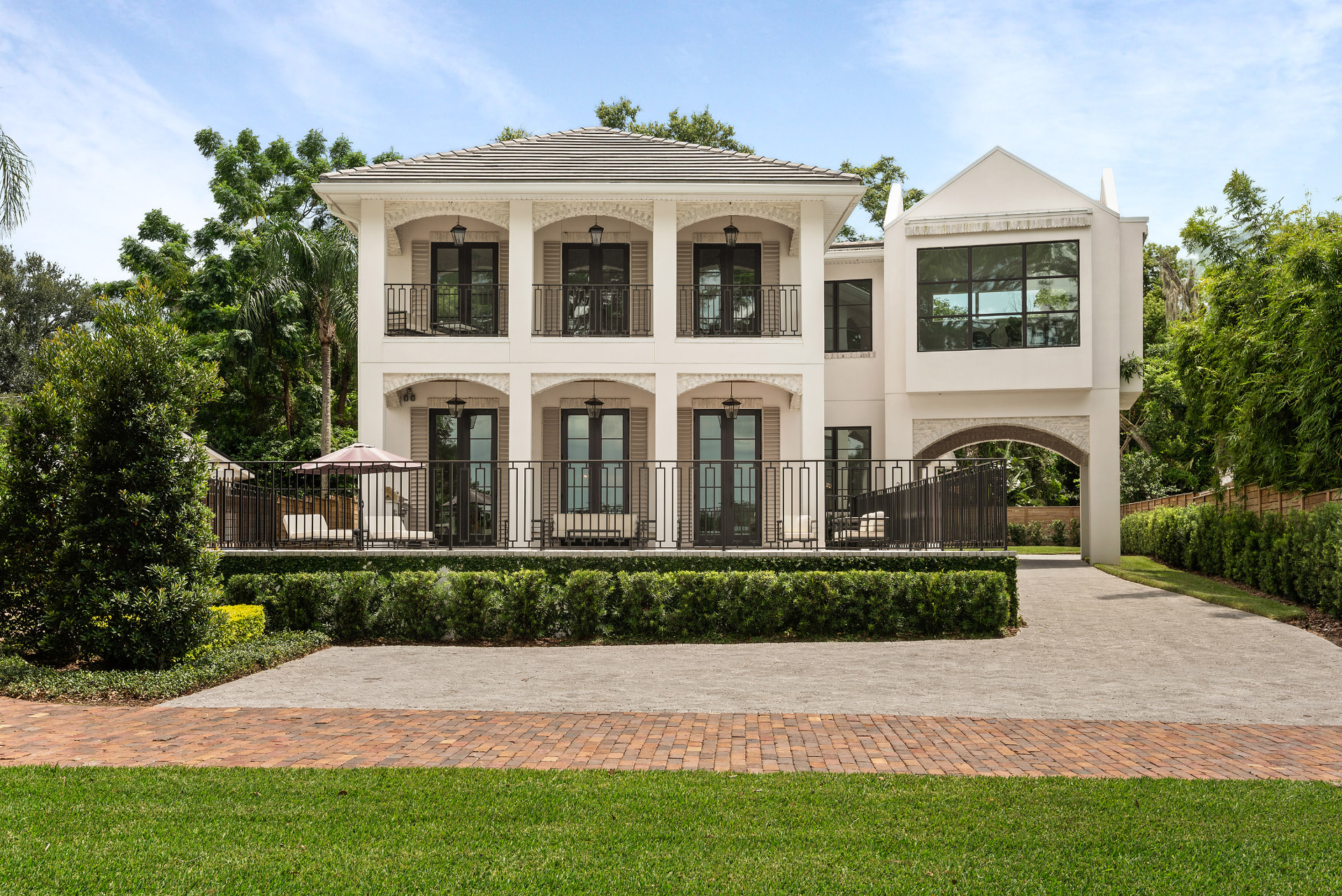 Elevation of Killarney: a two-storey cream stucco residence with arched openings on both levels, iron balcony railings, a terrace with lounge seating and a brick and paver drive.