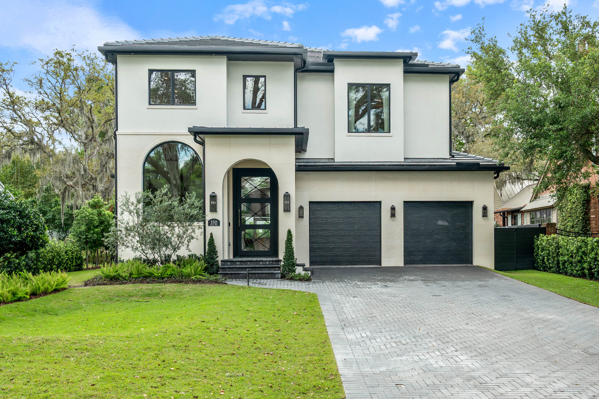 Front elevation of Dana: a two-storey white stucco home with a dark tile roof, arched entry with a black glass door, a tall arched window and two dark garage doors beside a herringbone paver driveway.