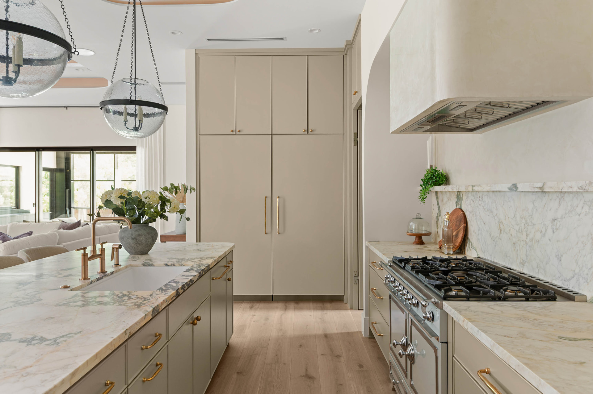 Kitchen at Dana with veined marble island and counters, brass fixtures, taupe cabinetry, a stainless range beneath a plaster hood and seeded glass globe pendants.