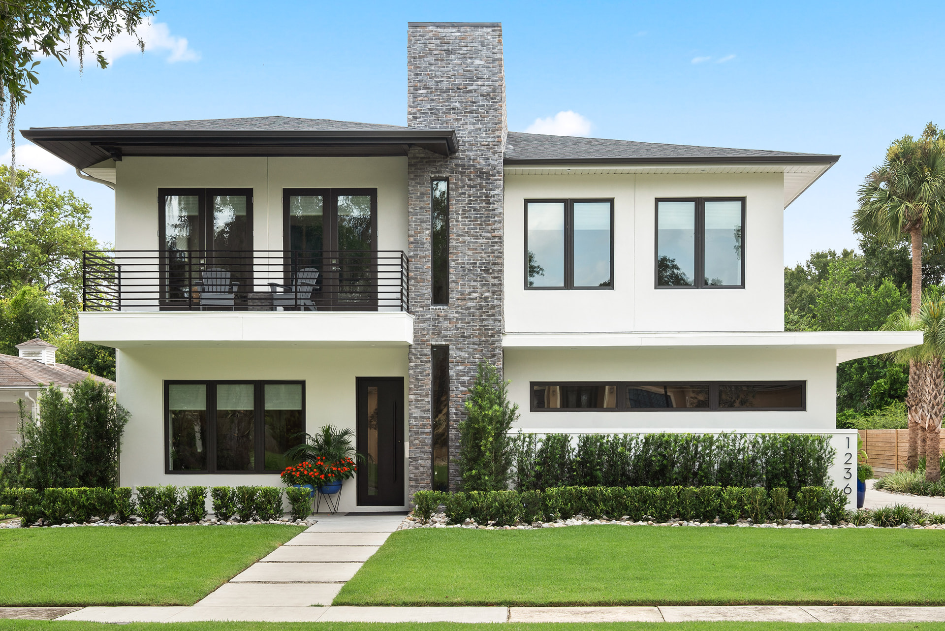 Front elevation of Nottingham: a prairie-style white stucco residence with wide overhanging eaves, a full-height grey brick chimney, a second-floor balcony with black metal railing and trimmed hedges along a concrete walk.