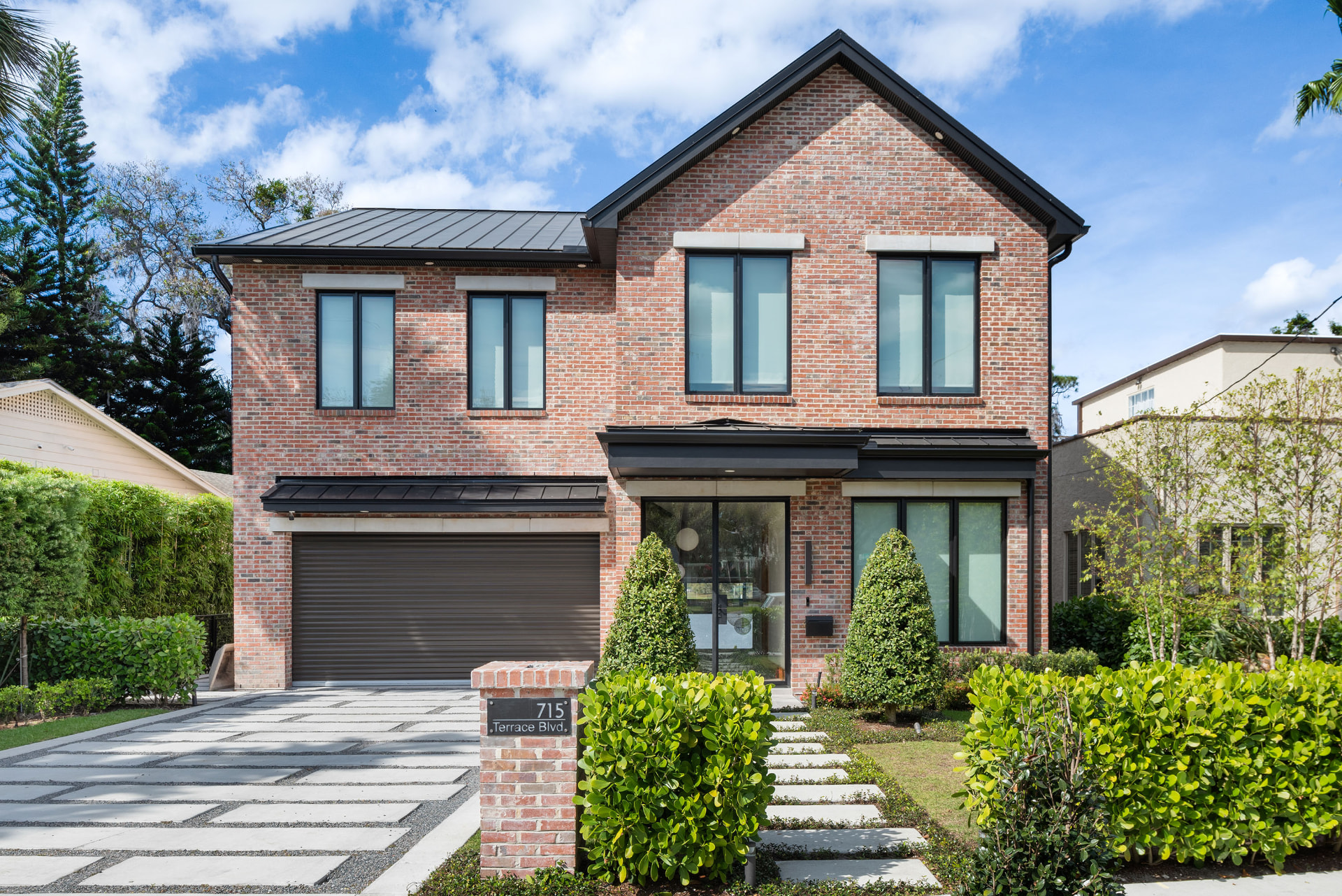 Front elevation of Terrace: a two-storey red brick residence with a steep gabled roof, black standing-seam awnings, black-framed windows and topiary shrubs beside a banded concrete drive.