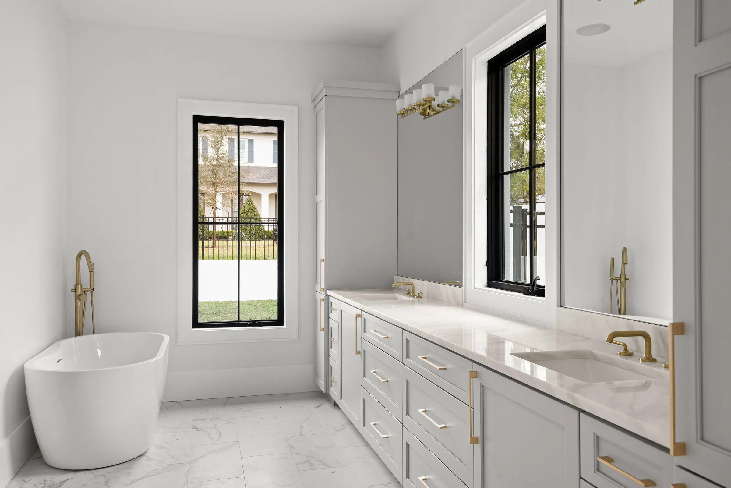 Bright bathroom with a freestanding tub, long double vanity, gold-toned fixtures and black-framed windows.