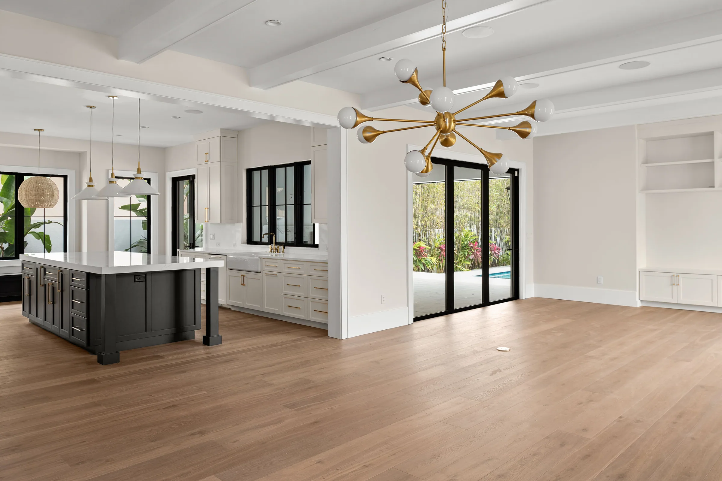 Open kitchen and living area with a charcoal island, pale floors, warm metallic lighting and rear glazing.