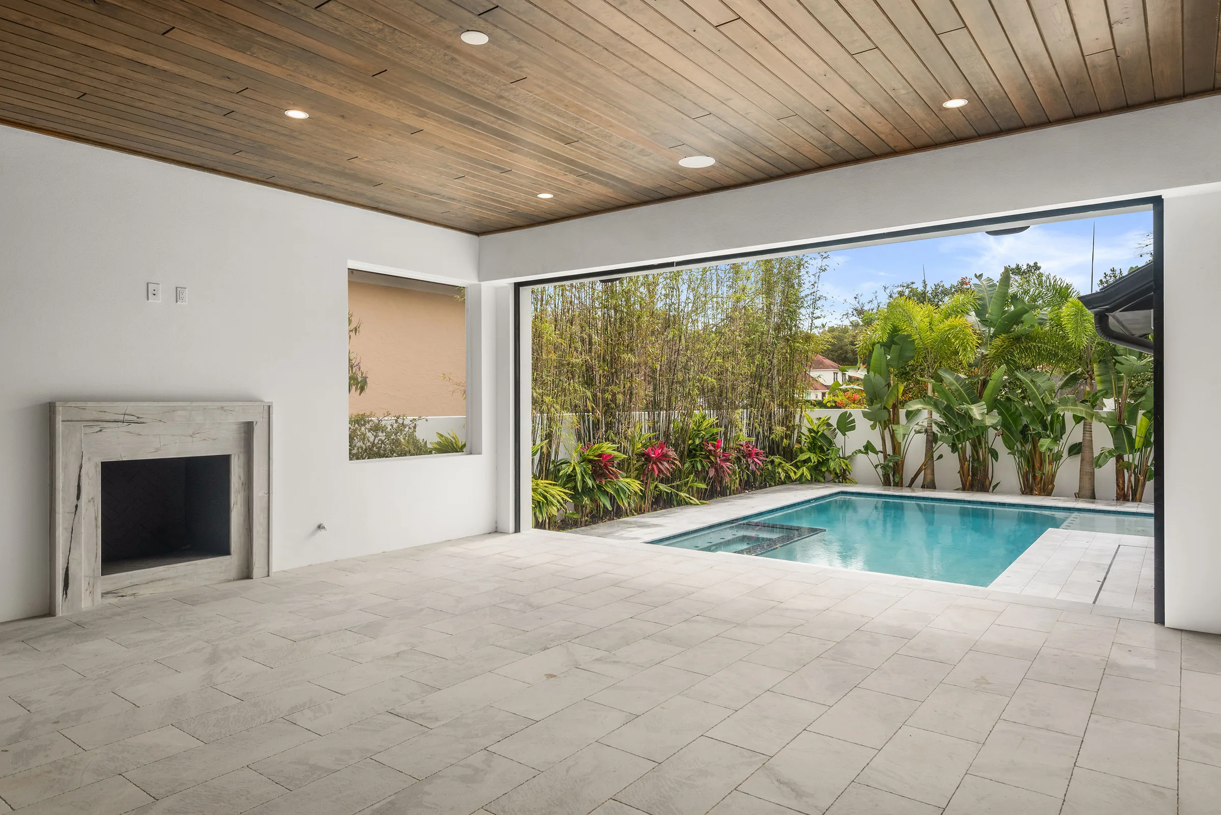 Covered lanai with a wood ceiling and fireplace opening to a pool edged by tropical planting.