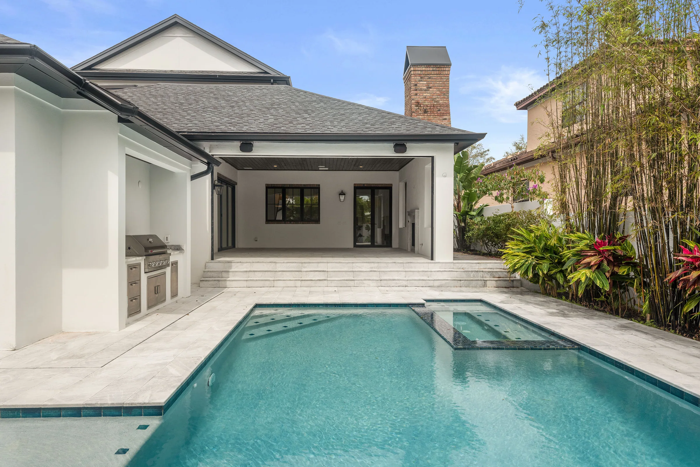 Pool and integrated spa beside a built-in grill, pale paving and a covered lanai with a brick chimney.