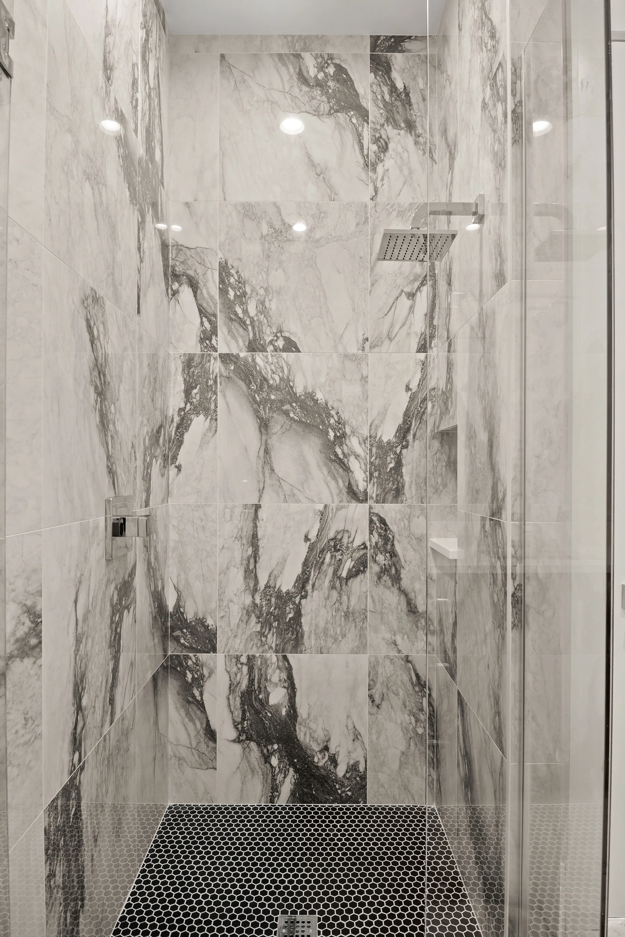 Glass-enclosed shower with strongly veined white-and-gray walls, a square showerhead and dark hexagonal floor tile.