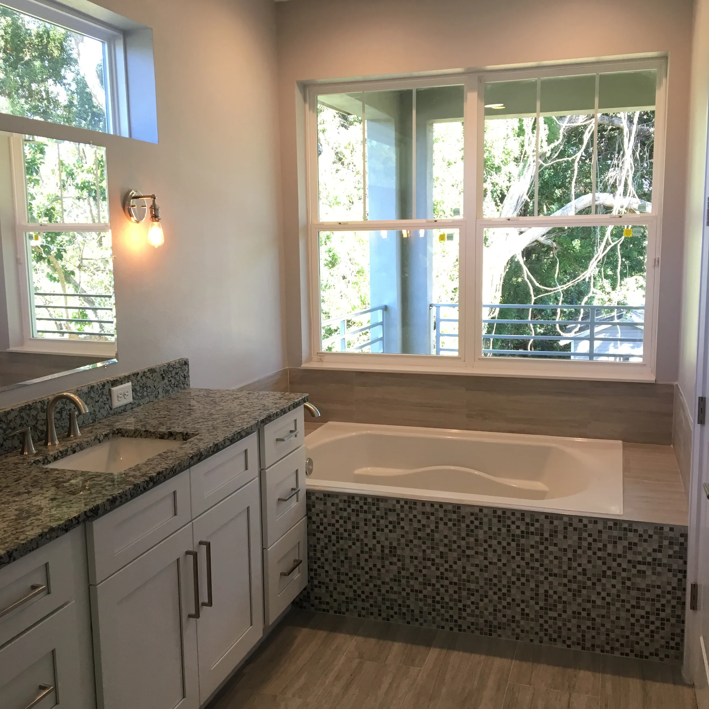 White vanity beside an inset bathtub with a mosaic-tile front beneath a large window.