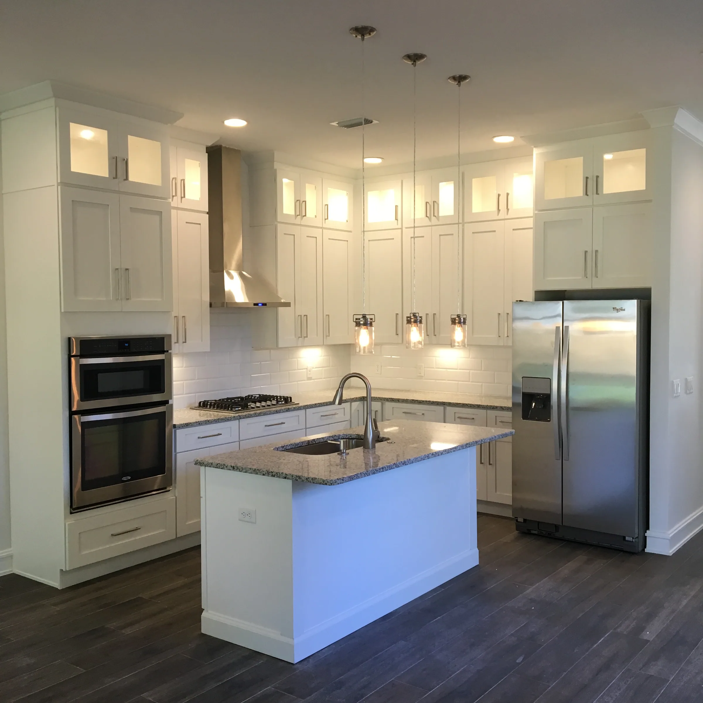 White kitchen with lit glass-front upper cabinets, an island sink and three glass pendant lights.