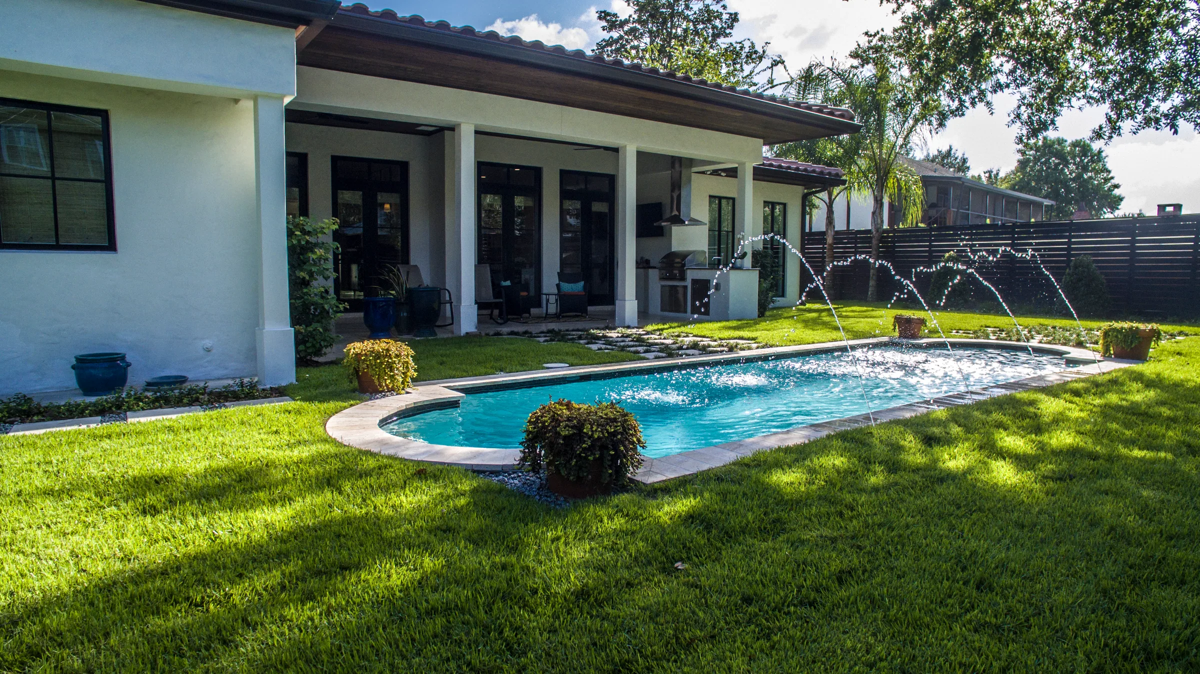 Garden pool with arcing water jets beside a covered terrace, outdoor grill and lawn.