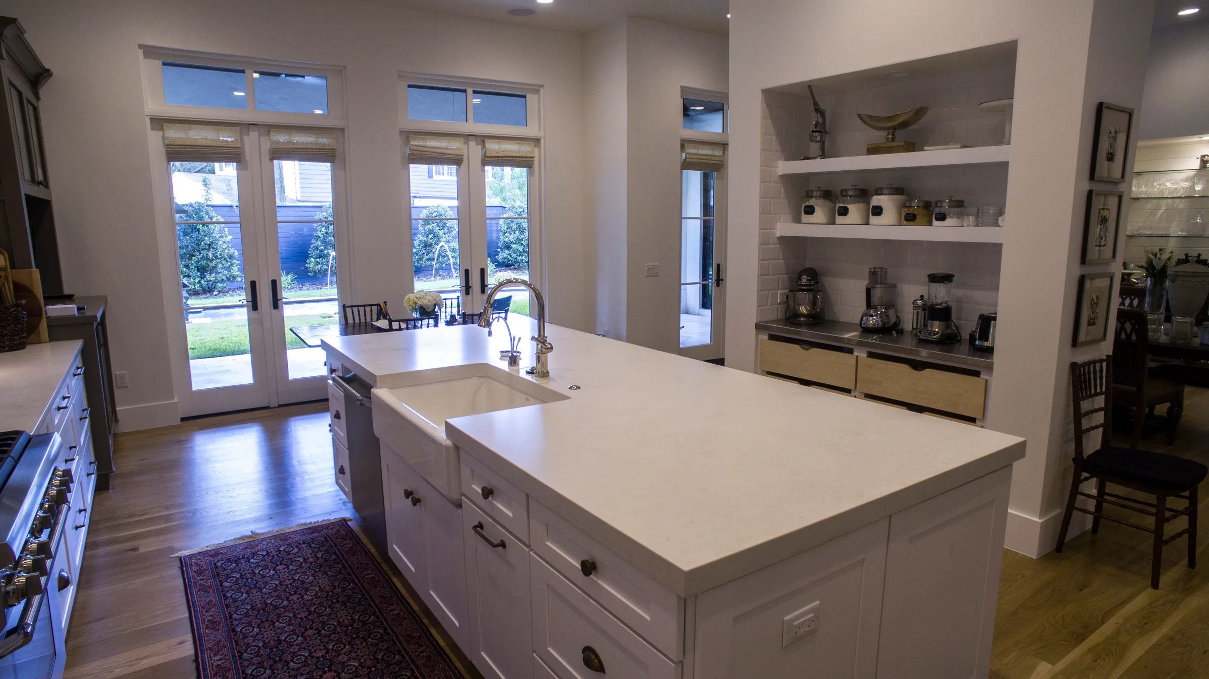 White kitchen island with an apron-front sink, beside a recessed appliance alcove and glazed doors overlooking the pool.