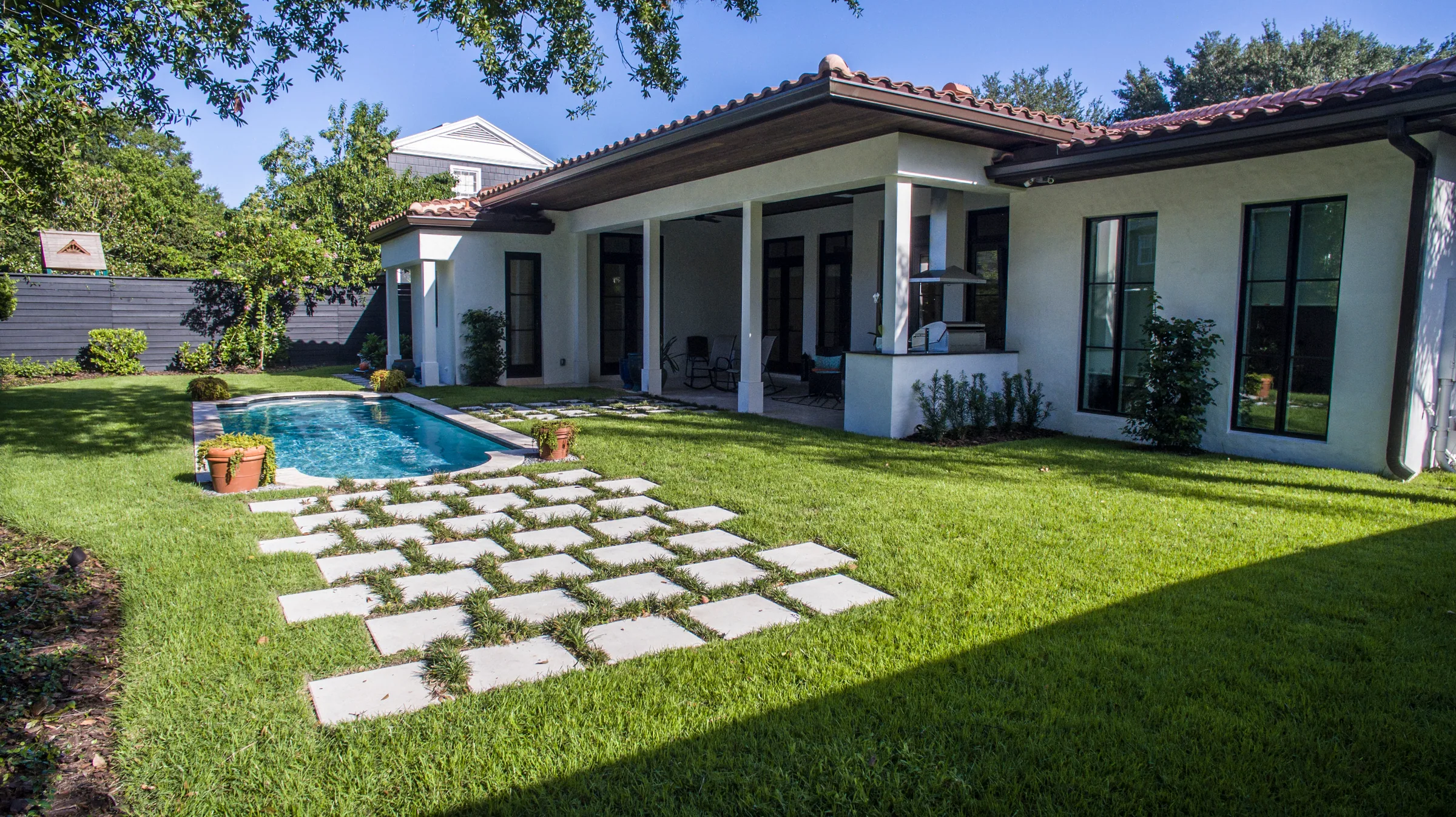 Covered rear terrace beside a garden pool, with square stepping stones set in a planted grid across the lawn.