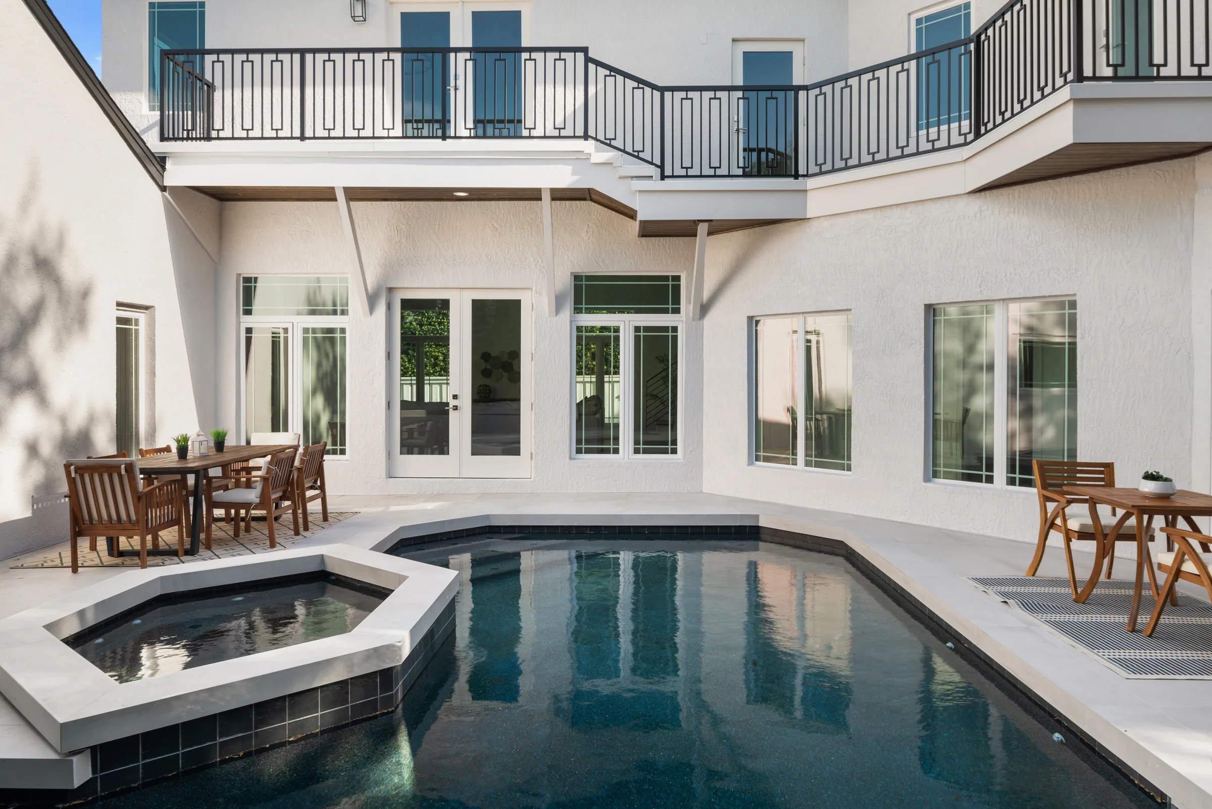 Rear courtyard with a rectangular pool, raised spa, paved terrace and upper balcony along the white residence.