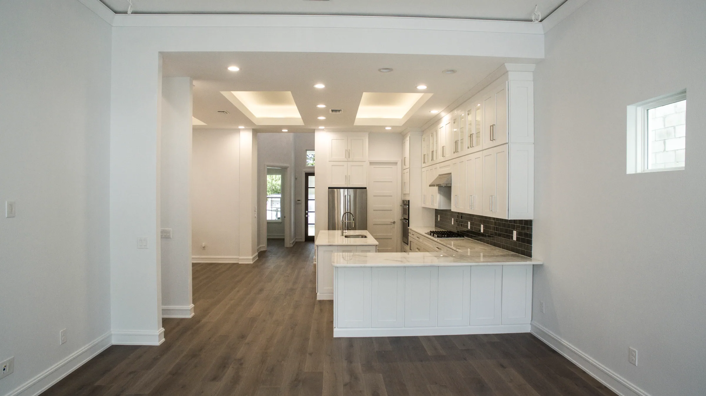 Long unfurnished living space opening to a white island kitchen beneath recessed ceiling details.