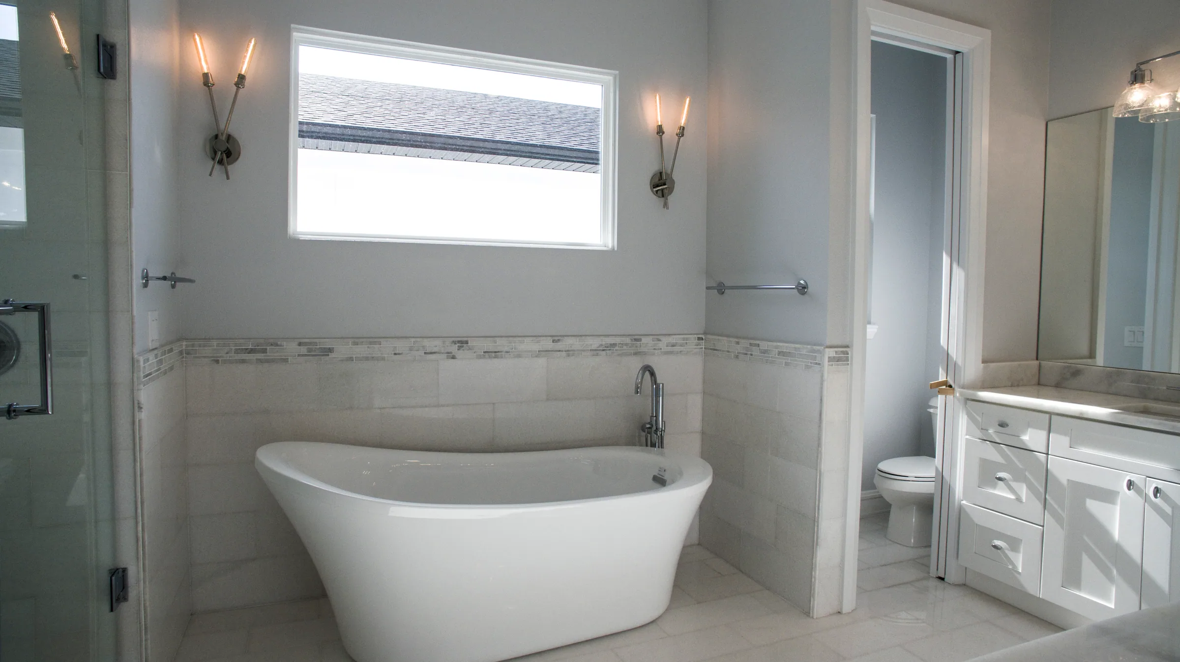 Bright bathroom with a freestanding white tub, glass-enclosed shower and long white vanity.
