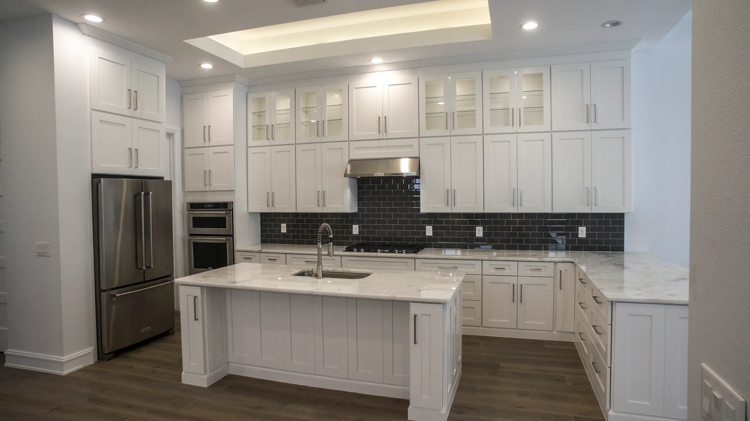 White kitchen with stacked upper cabinets, a pale island, dark tile backsplash and stainless appliances.