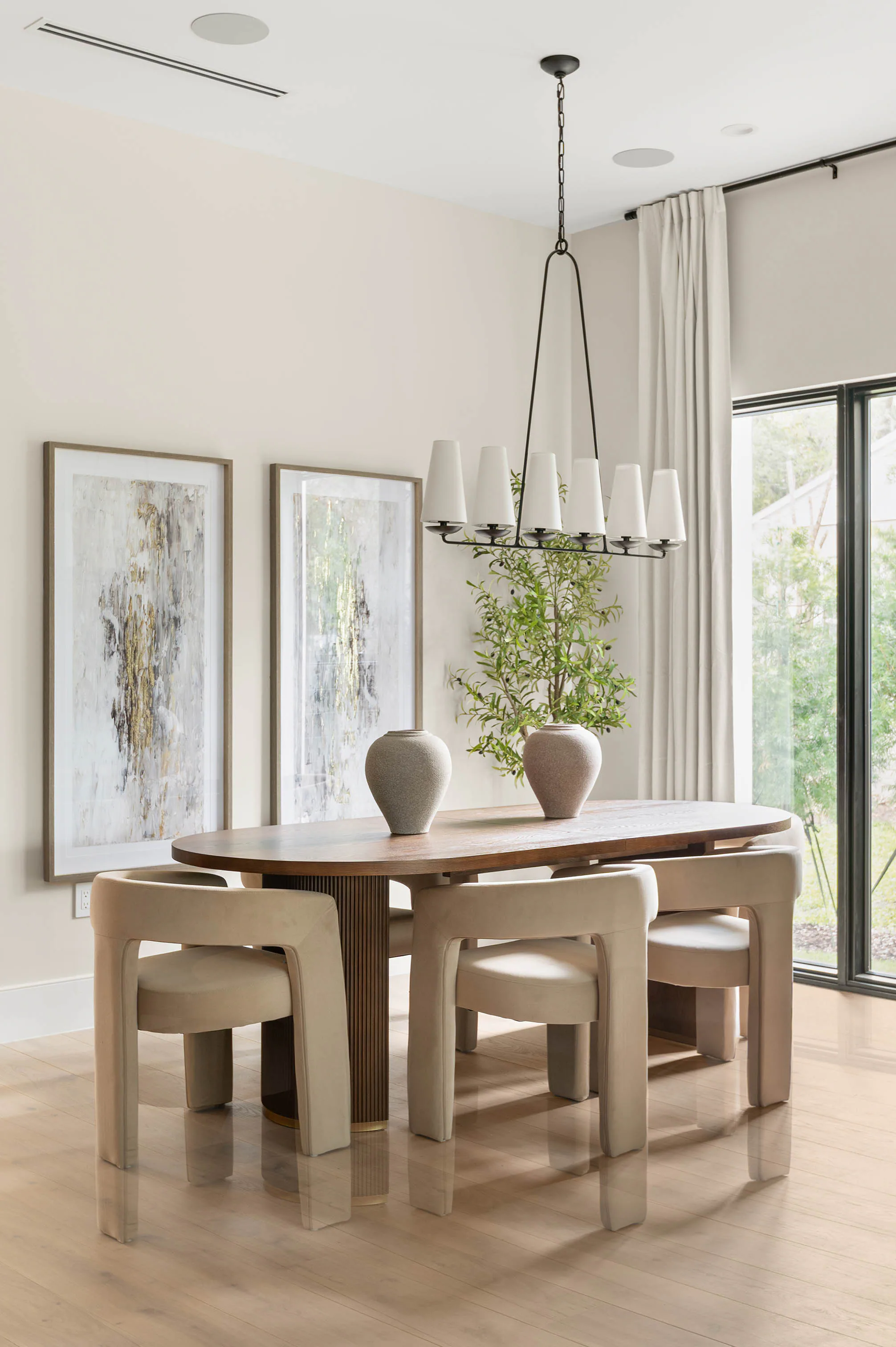 Oval dining table beside full-height windows and framed artwork.