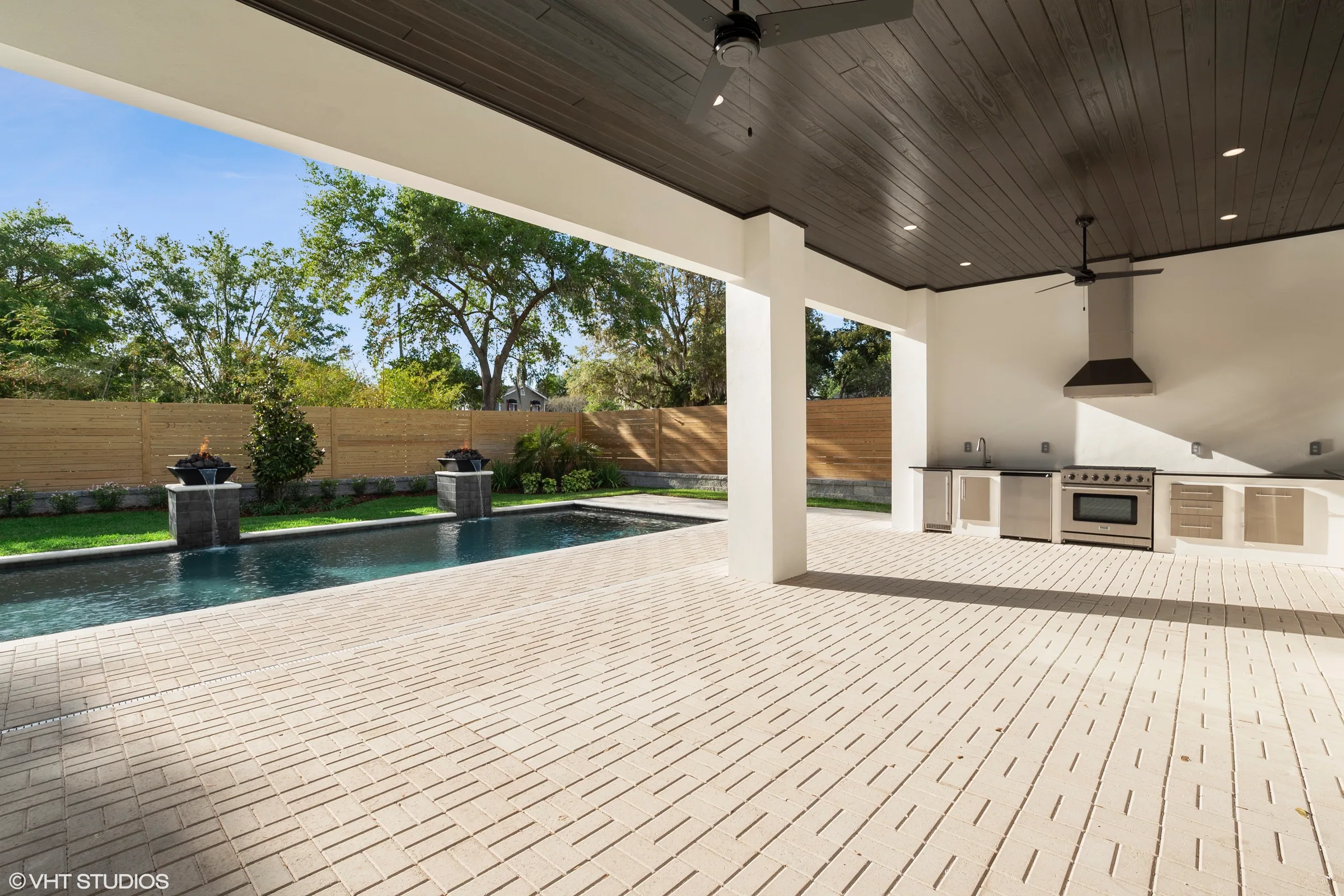 Covered patio with a dark paneled ceiling, fans and an outdoor cooking area beside a rectangular pool with fire-and-water bowls.
