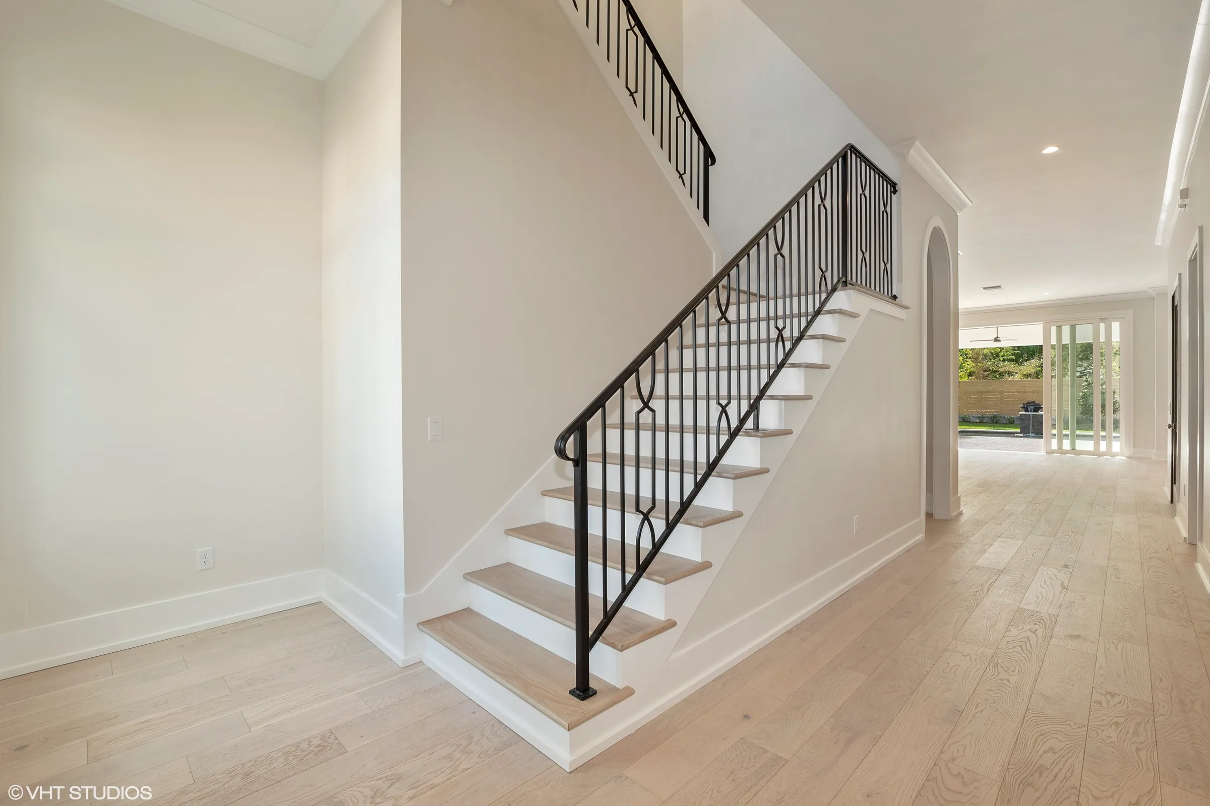 Foyer stair with dark geometric balusters and light-toned treads beside an arched passage leading toward the rear patio.
