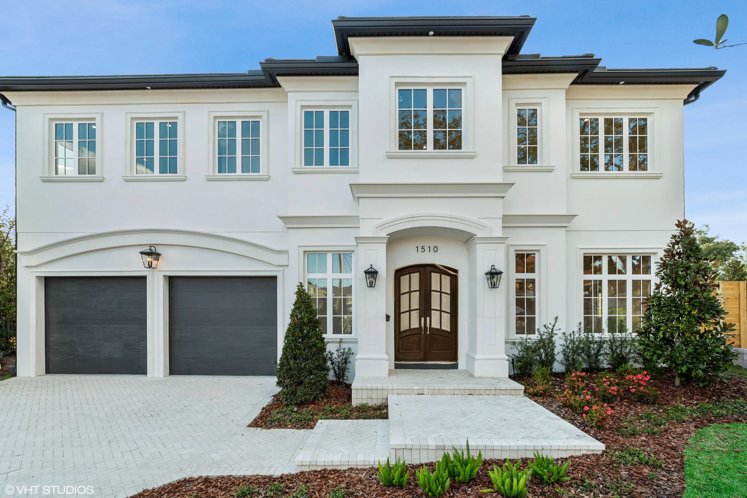 White two-story home with an arched double-door entry, dark garage doors, divided-light windows and a paver driveway.