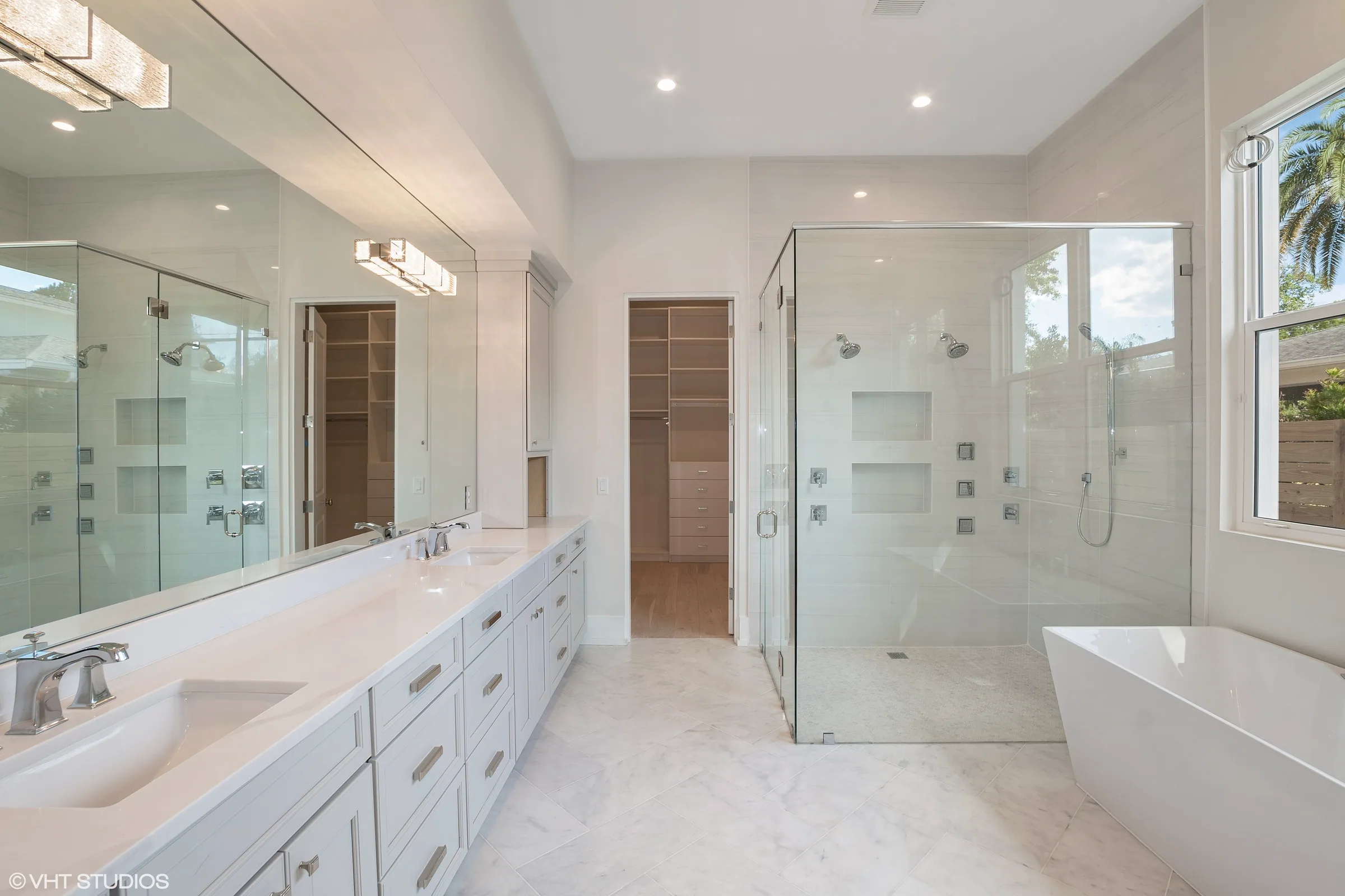 Pale bathroom with a long double vanity, wide mirror, glass-enclosed shower and freestanding tub beside a window.