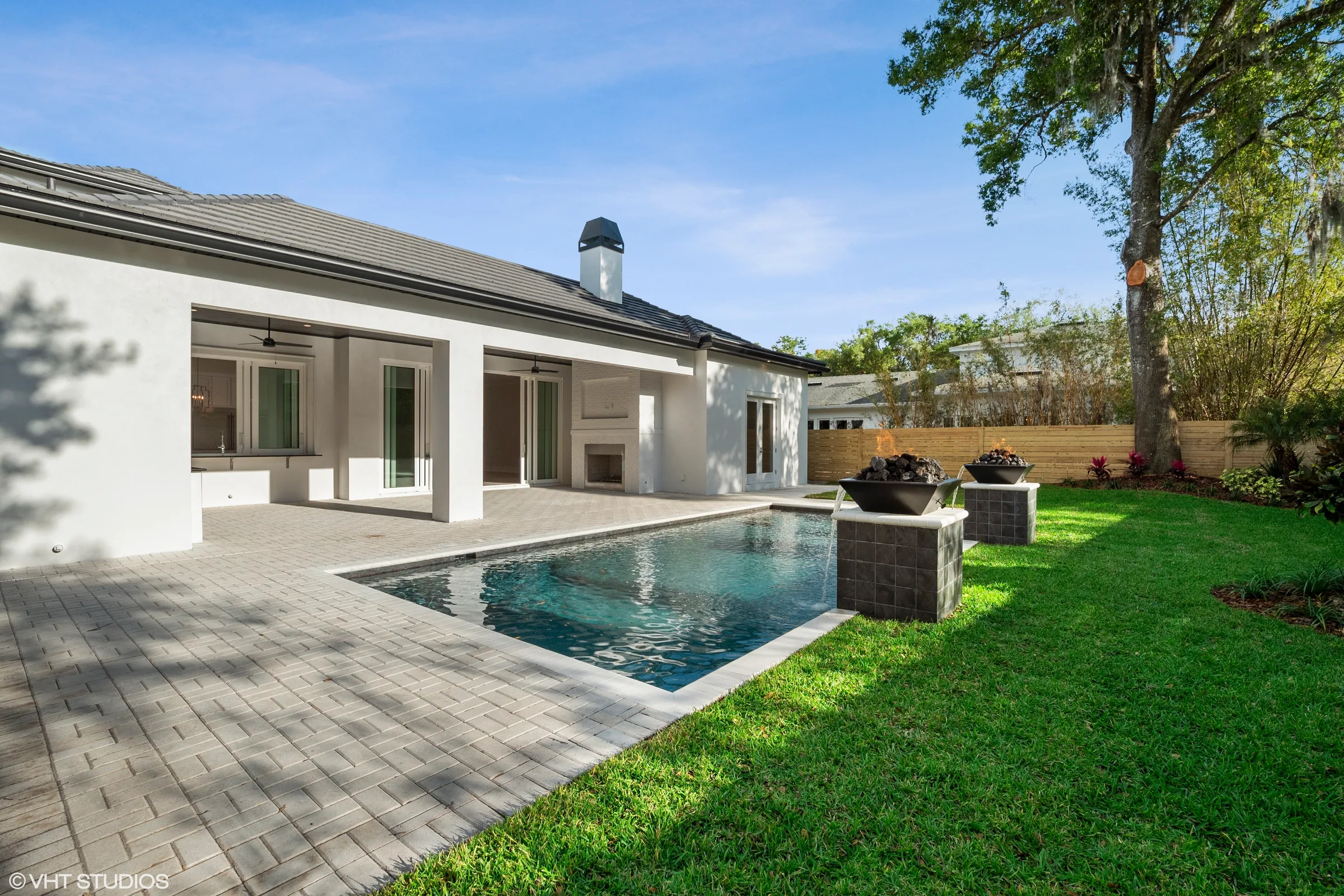 Rectangular pool with two fire-and-water bowls facing a covered rear patio with a fireplace, sliding doors and kitchen pass-through.