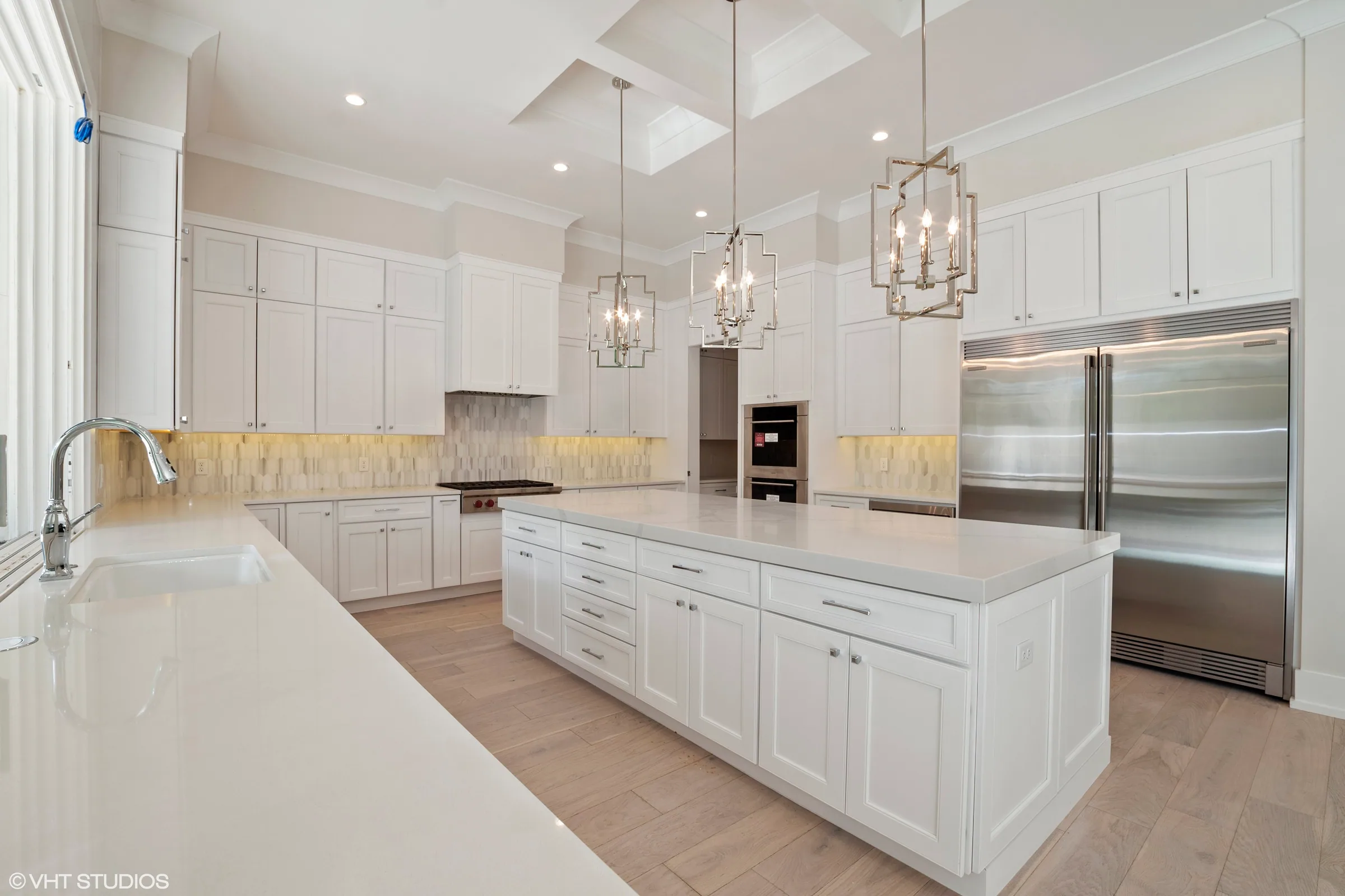 White kitchen with a large island, geometric pendant lights, recessed ceiling panels and a pale patterned backsplash.