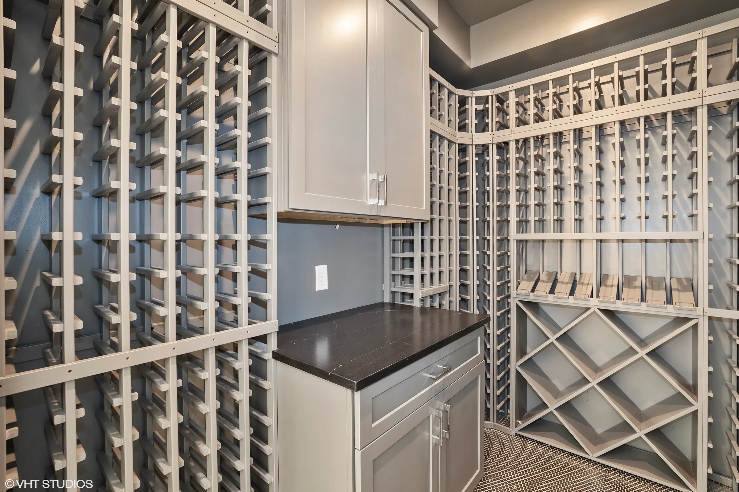 Wine storage room with built-in bottle racks, diagonal lower bins, gray cabinetry and a dark countertop.
