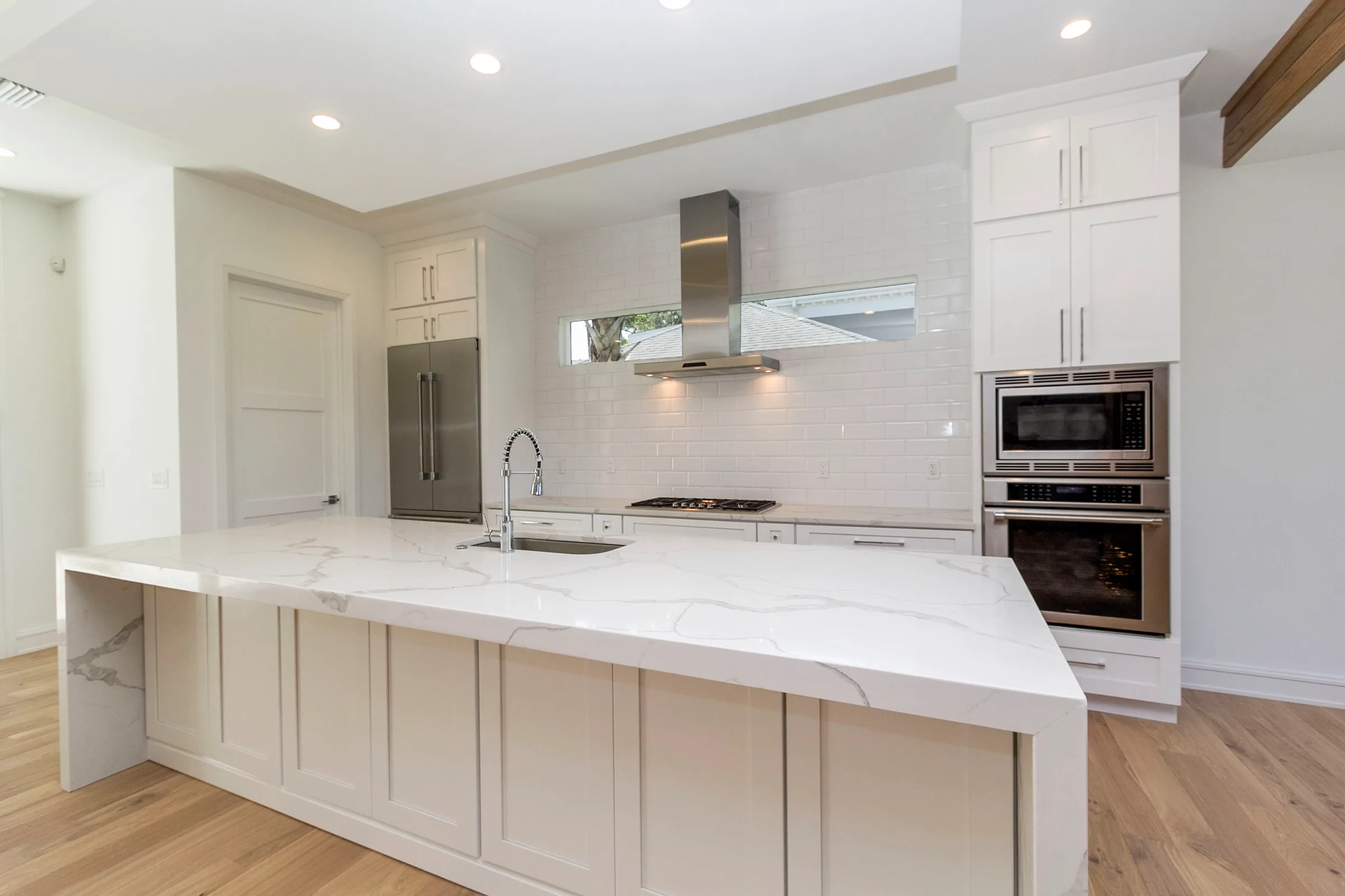 Wilbar kitchen with white cabinetry, a long island and sink, stainless hood and built-in ovens.