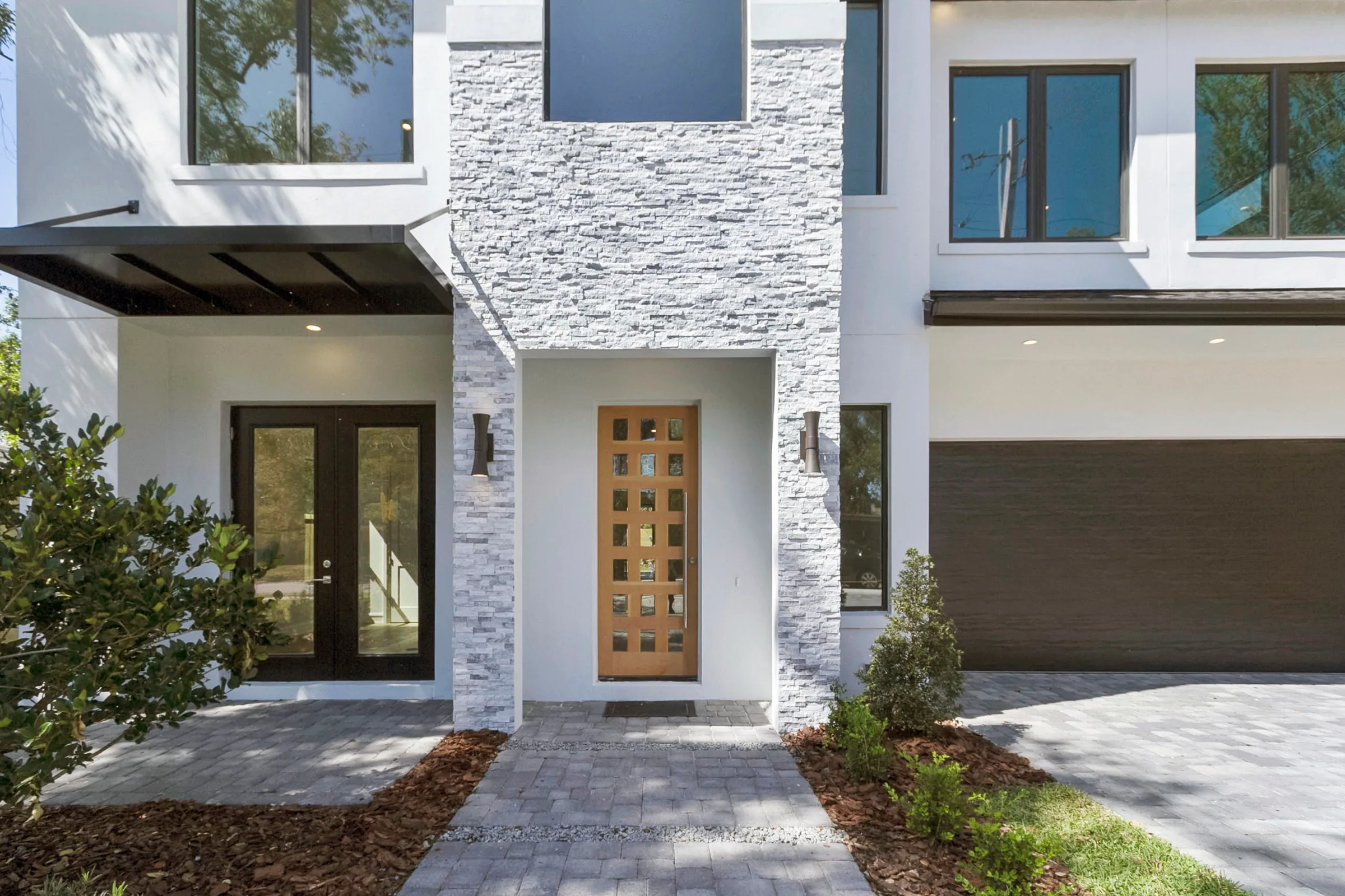 Glazed wood-toned front door framed by light textured cladding, dark windows and a paved approach.