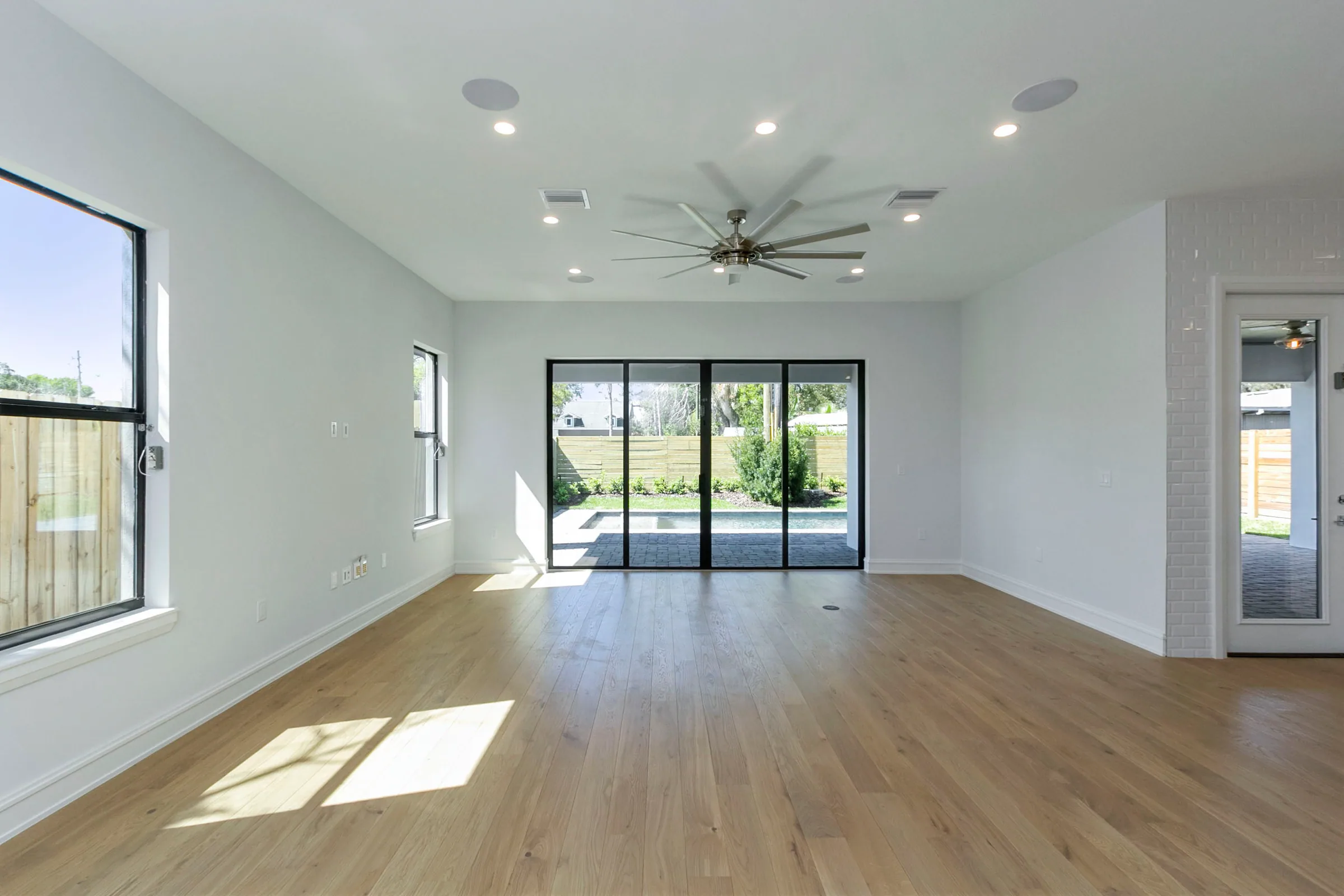 Unfurnished living room with pale wood-look flooring, dark-framed windows and sliding doors overlooking the pool.