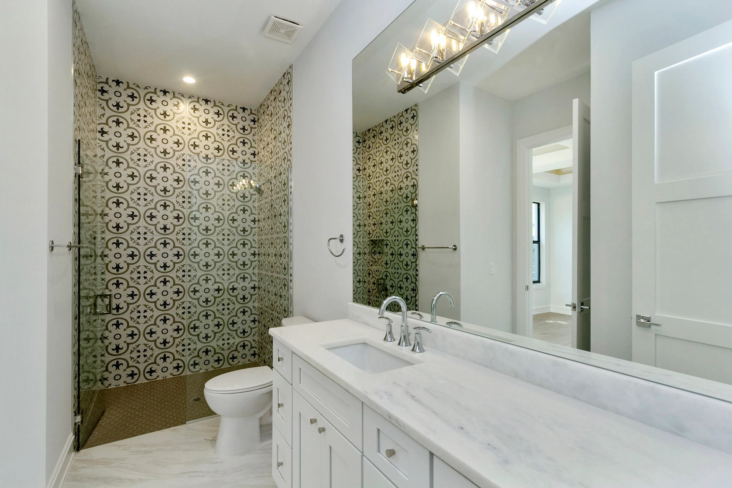 Glass shower with patterned wall tile beside a white vanity, broad mirror and chrome faucet.