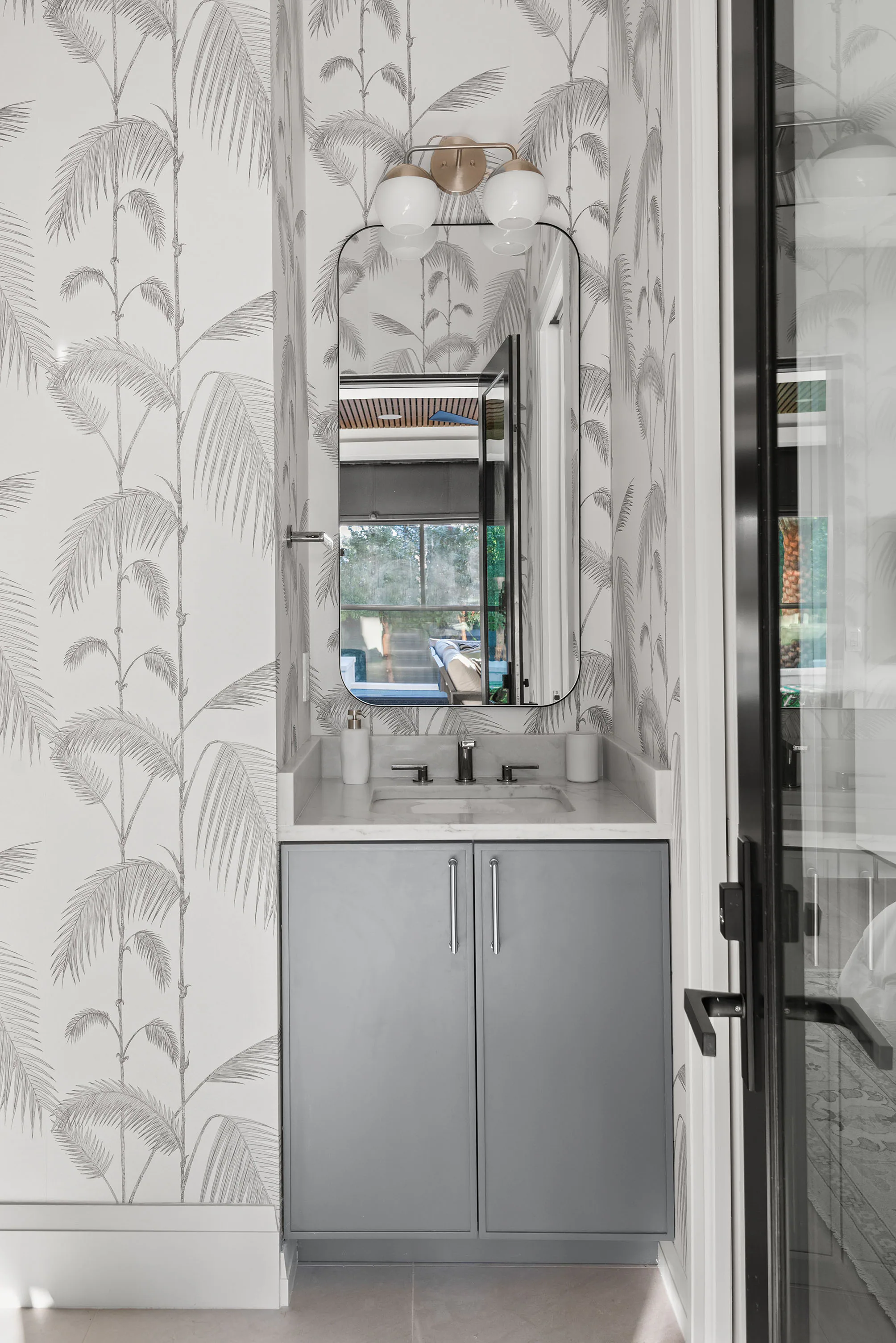 Powder room with palm-patterned wallpaper, a grey vanity and glass shower door. Rockwood, a custom home by Rare Builders in Orlando.