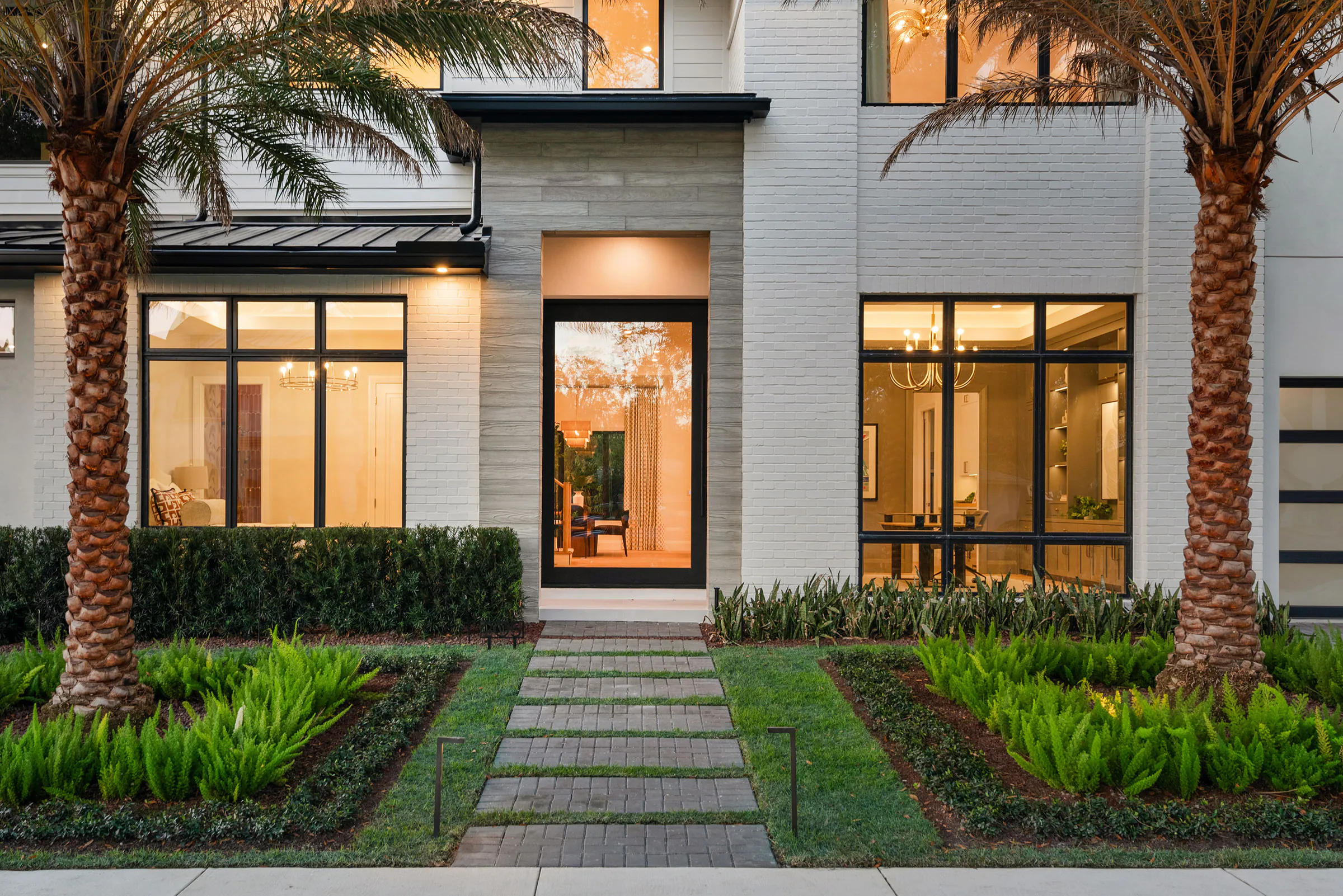 Rear entry glowing at dusk behind clipped hedges and palms.