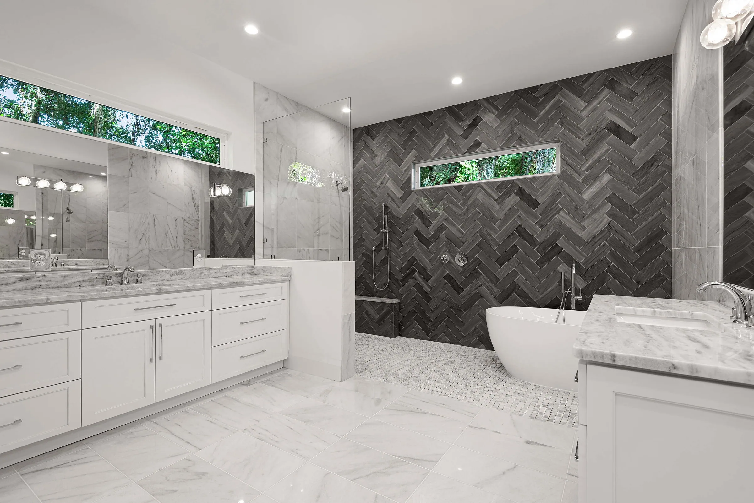 White bathroom with a dark herringbone-tile wall, freestanding tub, glass shower partition and broad vanity.