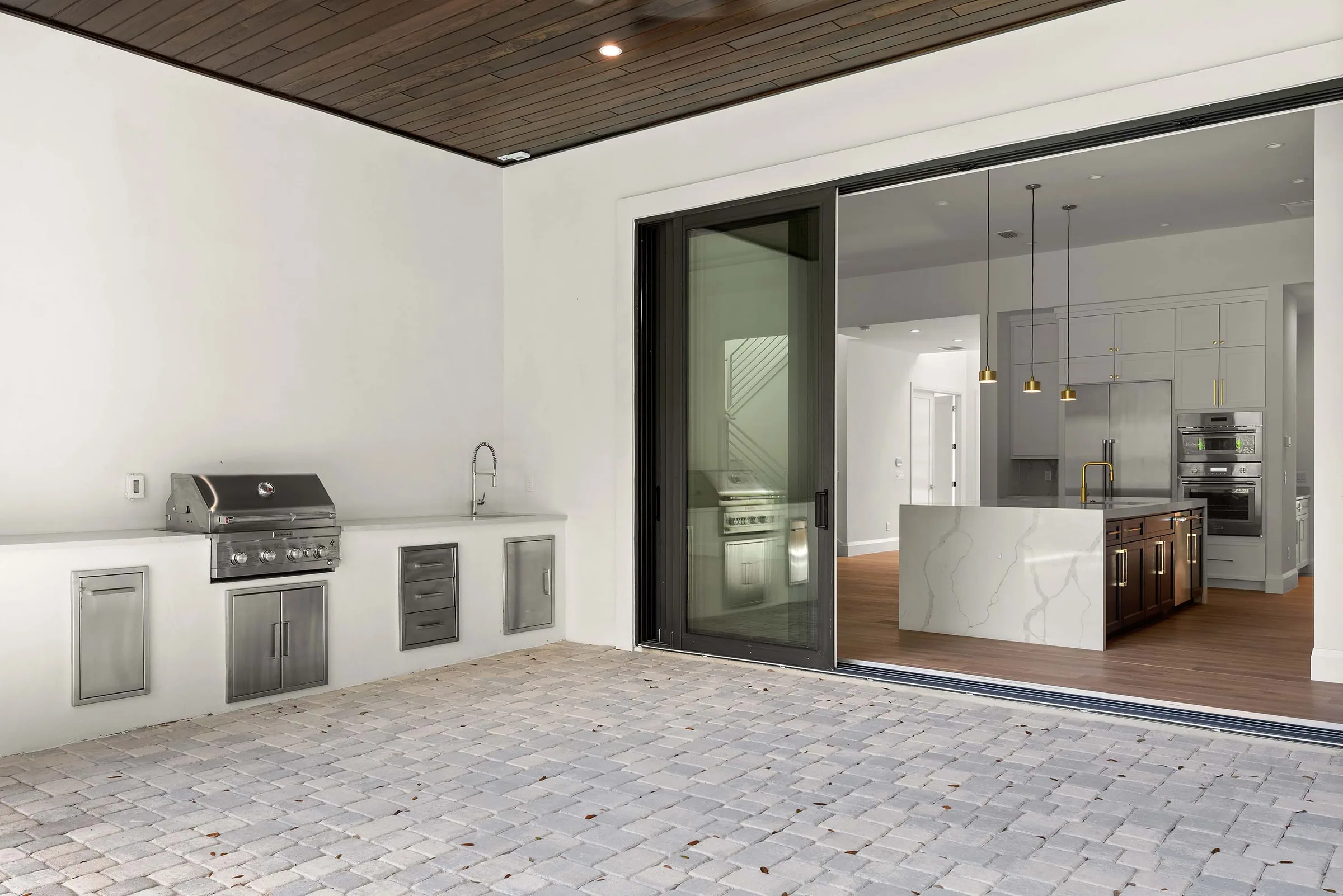 Covered paver patio with a built-in grill and sink beside open sliding doors to the kitchen.