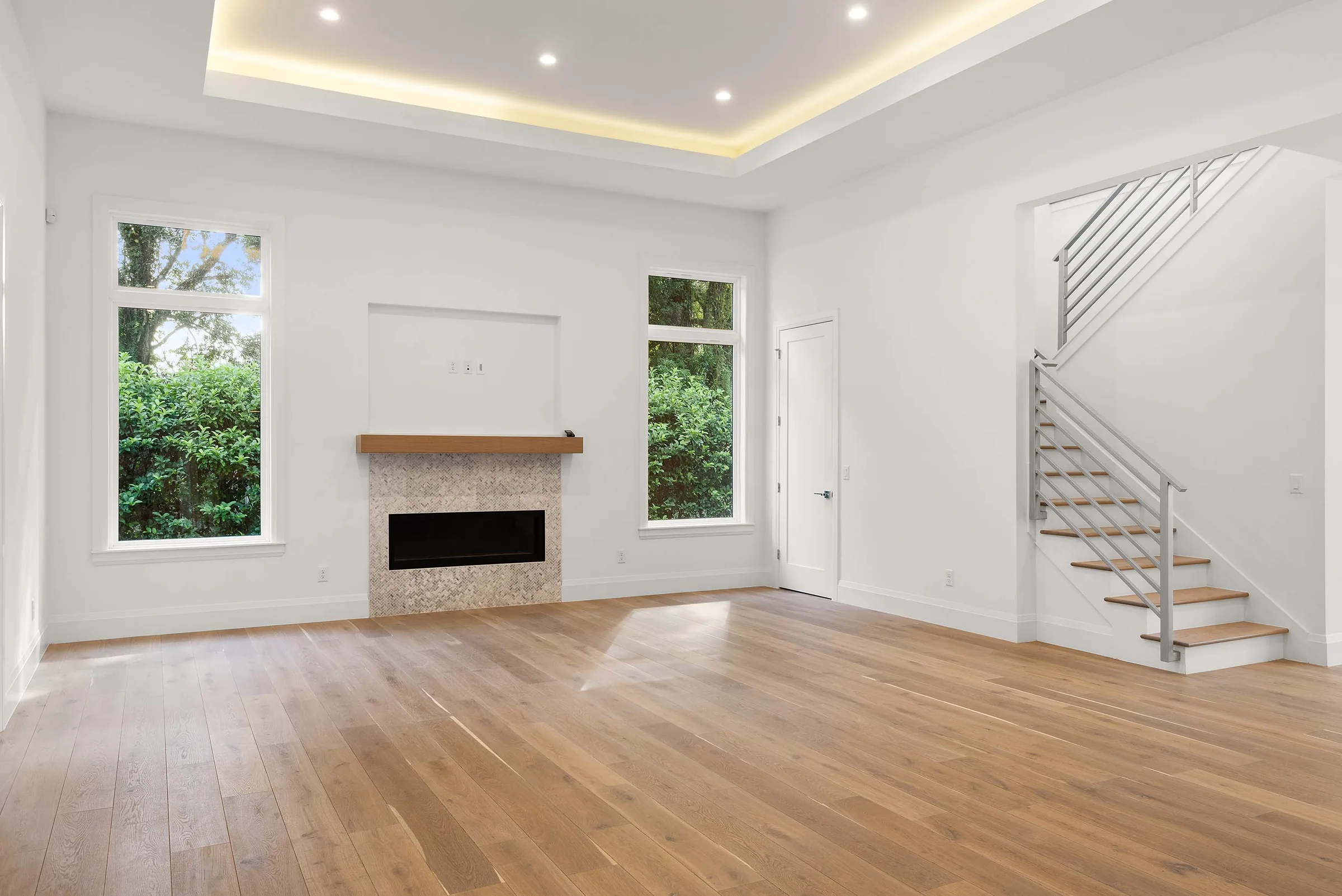 Living room with a tiled fireplace, wood-toned mantel, illuminated tray ceiling and open stair with horizontal railings.