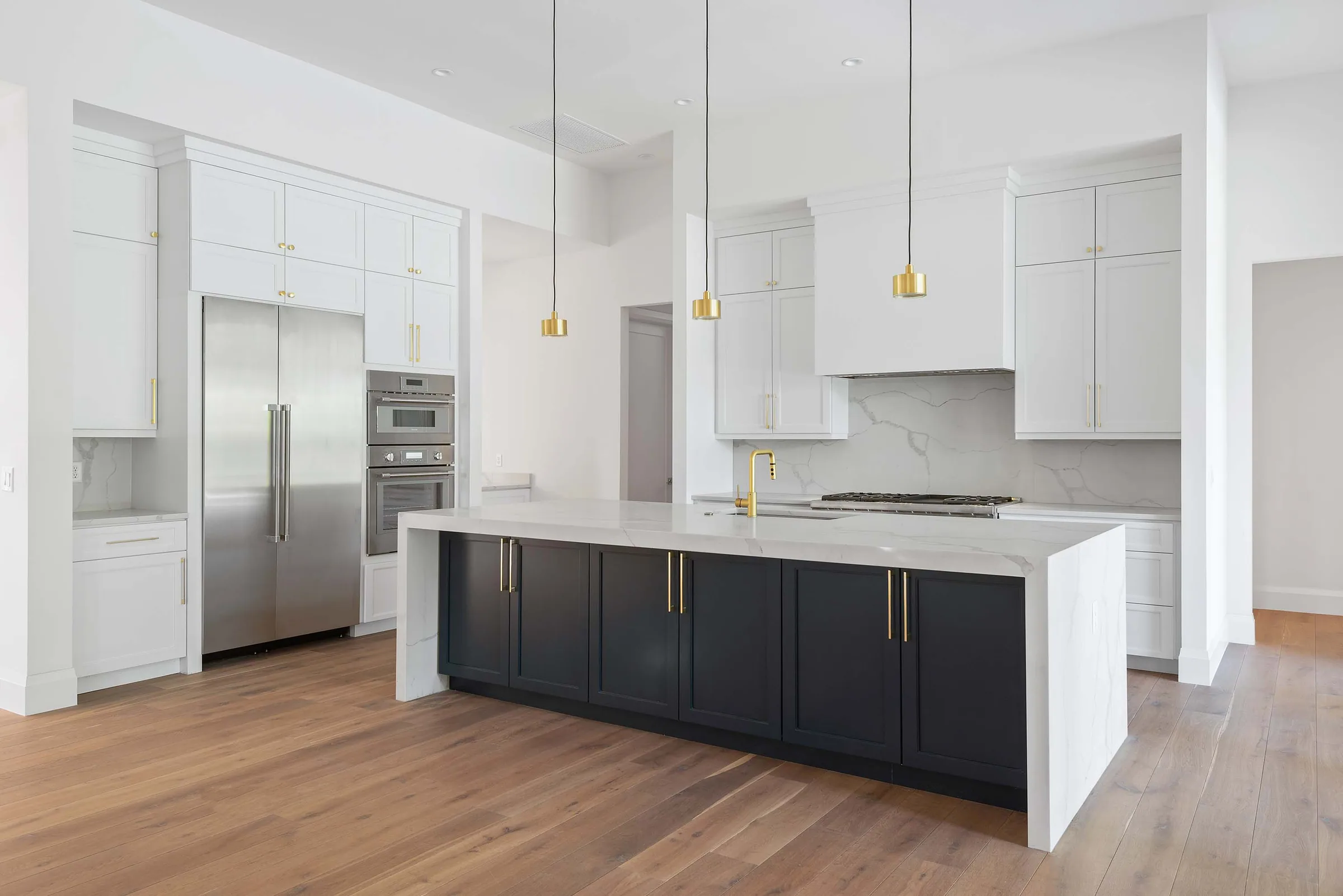 White kitchen with a navy island, pale waterfall countertop and three small gold-toned pendant lights.