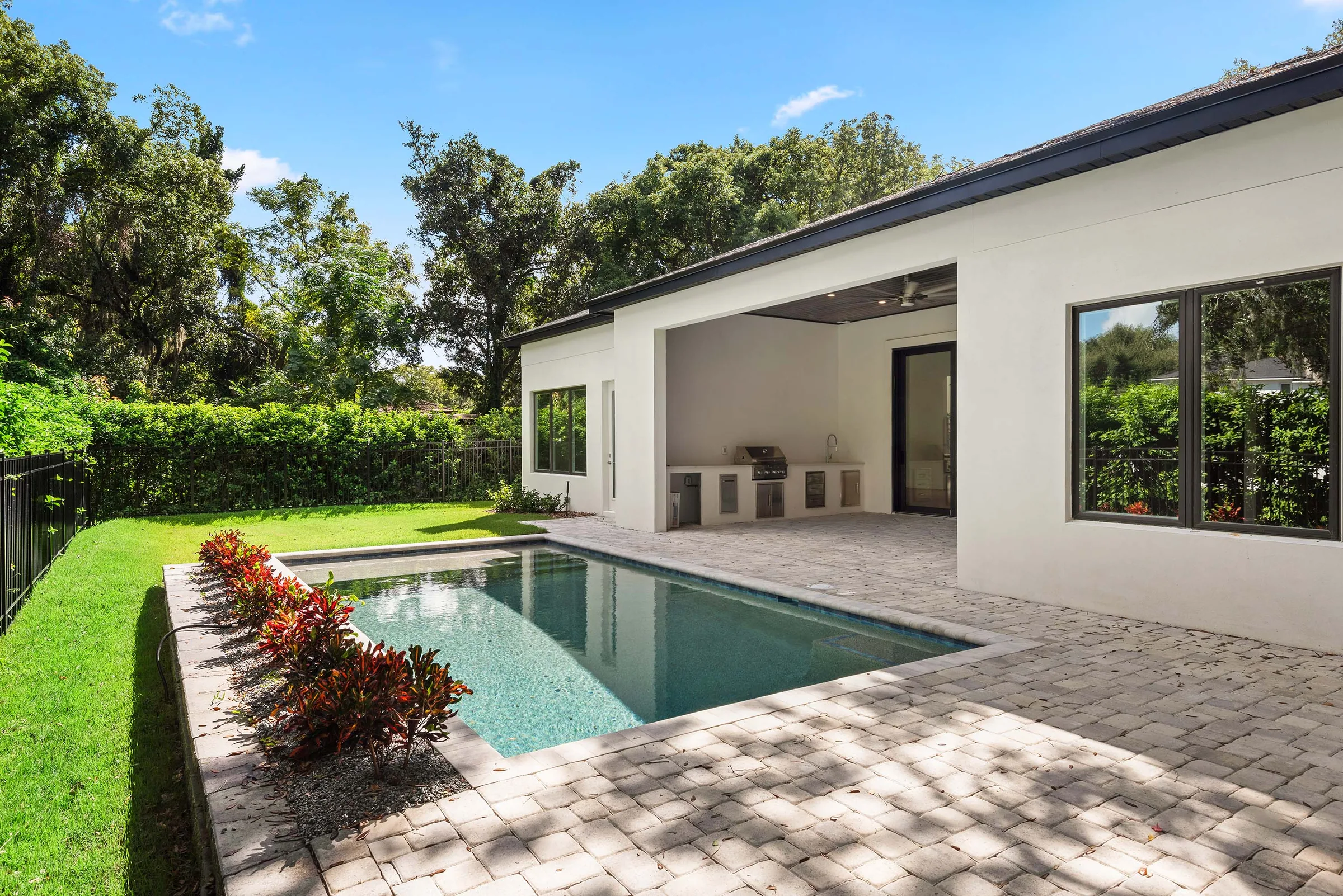 Rectangular pool beside a paver terrace, covered summer kitchen, lawn and planted boundary.