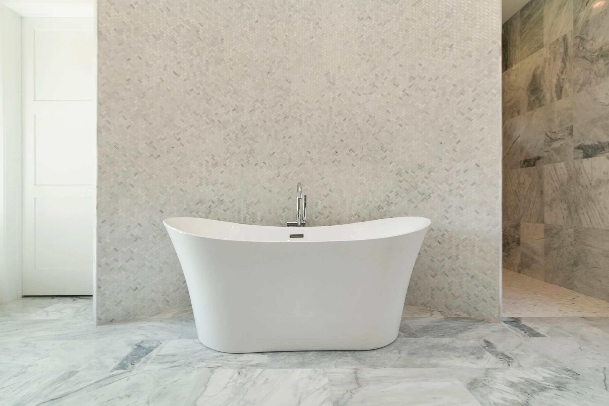 White freestanding bathtub in front of a pale herringbone-tile wall beside an open shower.