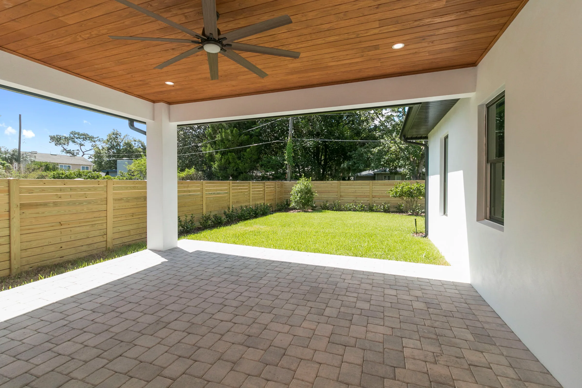 Covered paver porch with a wood-plank ceiling and large fan overlooking a fenced rear lawn.