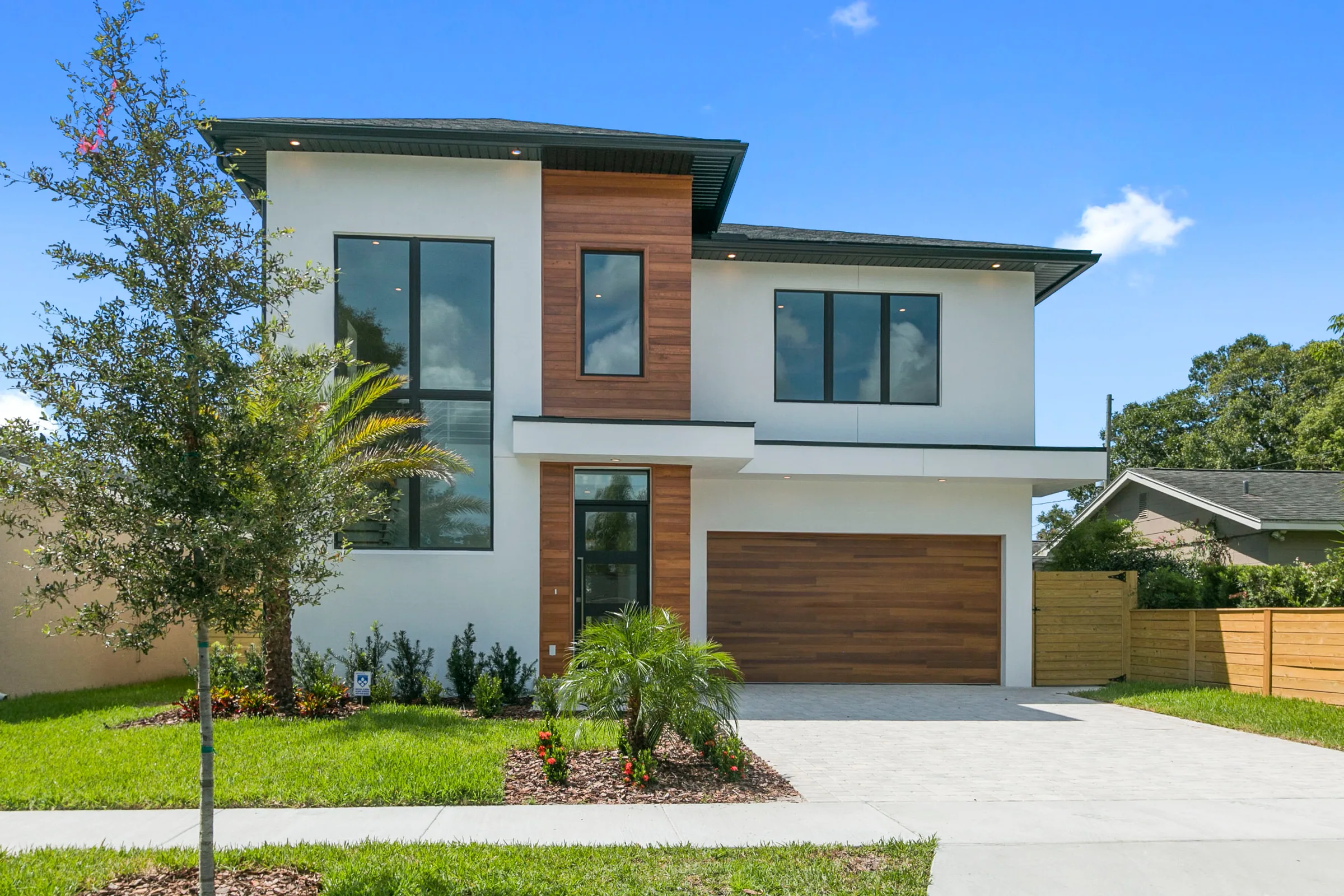 Daylight view of a two-story stucco home with wood-toned accents, dark-framed windows and a landscaped front yard.
