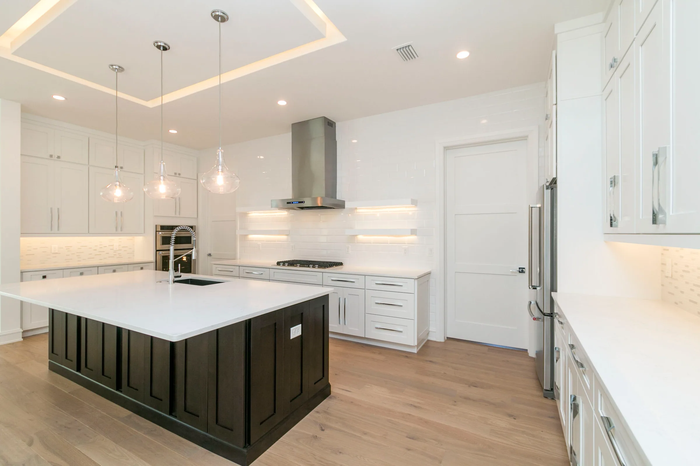 Dark kitchen island with white cabinetry, glass pendant lights and illuminated floating shelves beside a stainless range hood.