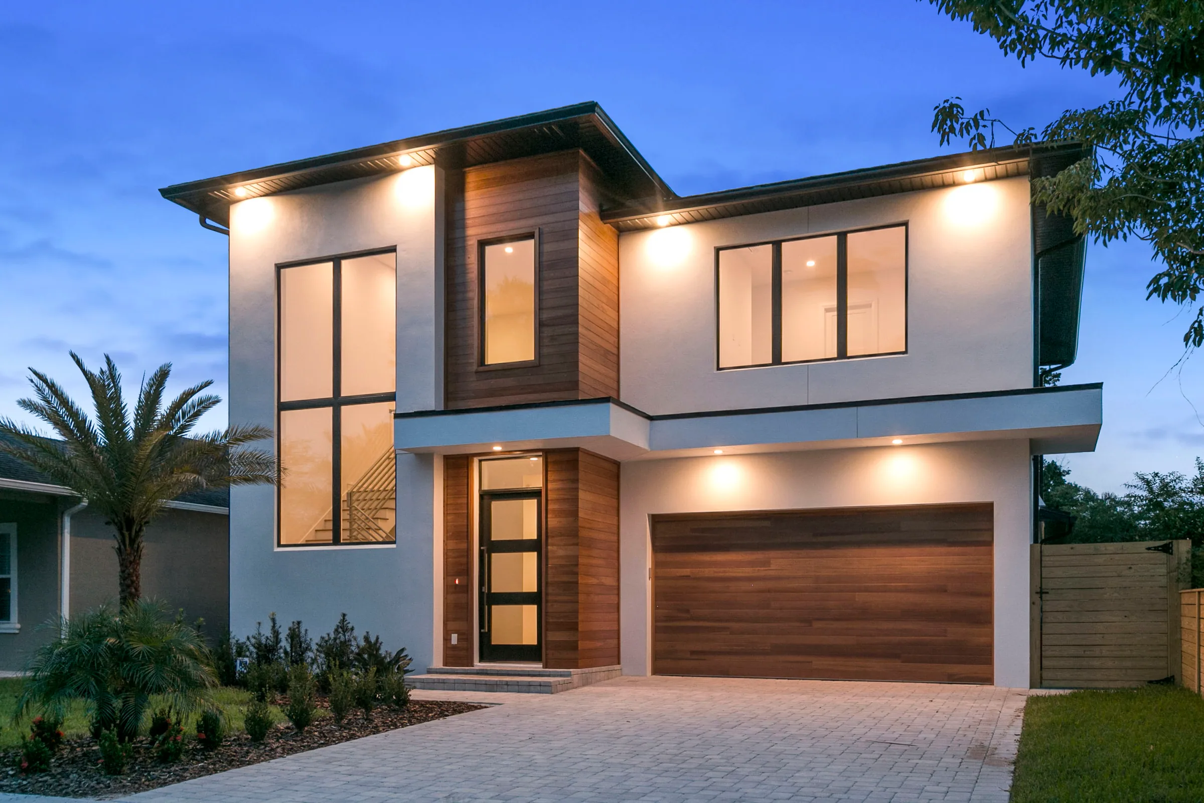 Two-story home at dusk with a tall stair window, wood-toned entry accents and an illuminated paver driveway.