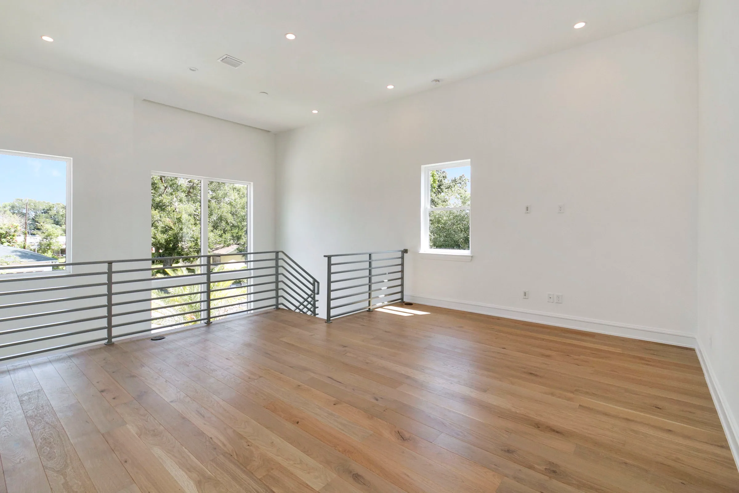 Open upper landing with pale plank flooring and horizontal metal railings beside large front windows.