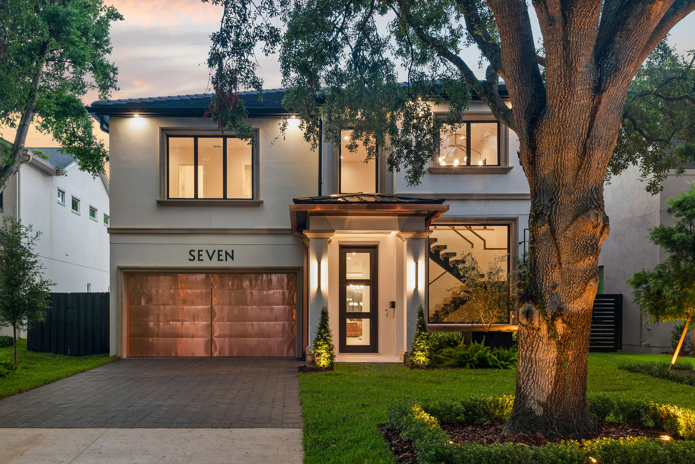 Front elevation glowing at dusk beneath mature oaks.